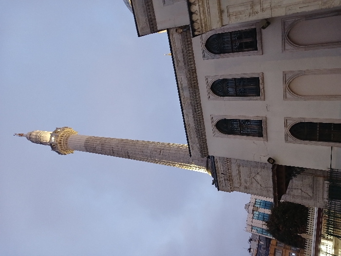 The image shows the exterior of the Süleymaniye Mosque in Istanbul, Türkiye, located at the coordinates 41.01, 28.953. The tall minaret of the mosque dominates the scene, rising up against a light blue sky. The minaret is a cylindrical structure with a series of balconies along its height. The mosque itself is a large, white building with arched windows and a stone facade. The mosque appears to be constructed with stone, while the minaret appears to be constructed with brick. The scene is captured in the late afternoon or early evening, with the soft light of the setting sun illuminating the mosque. The overall atmosphere is tranquil and serene. A low, white building with arched windows is in the foreground. Some trees are visible in the bottom left of the image, partially obscuring a taller building with windows.
