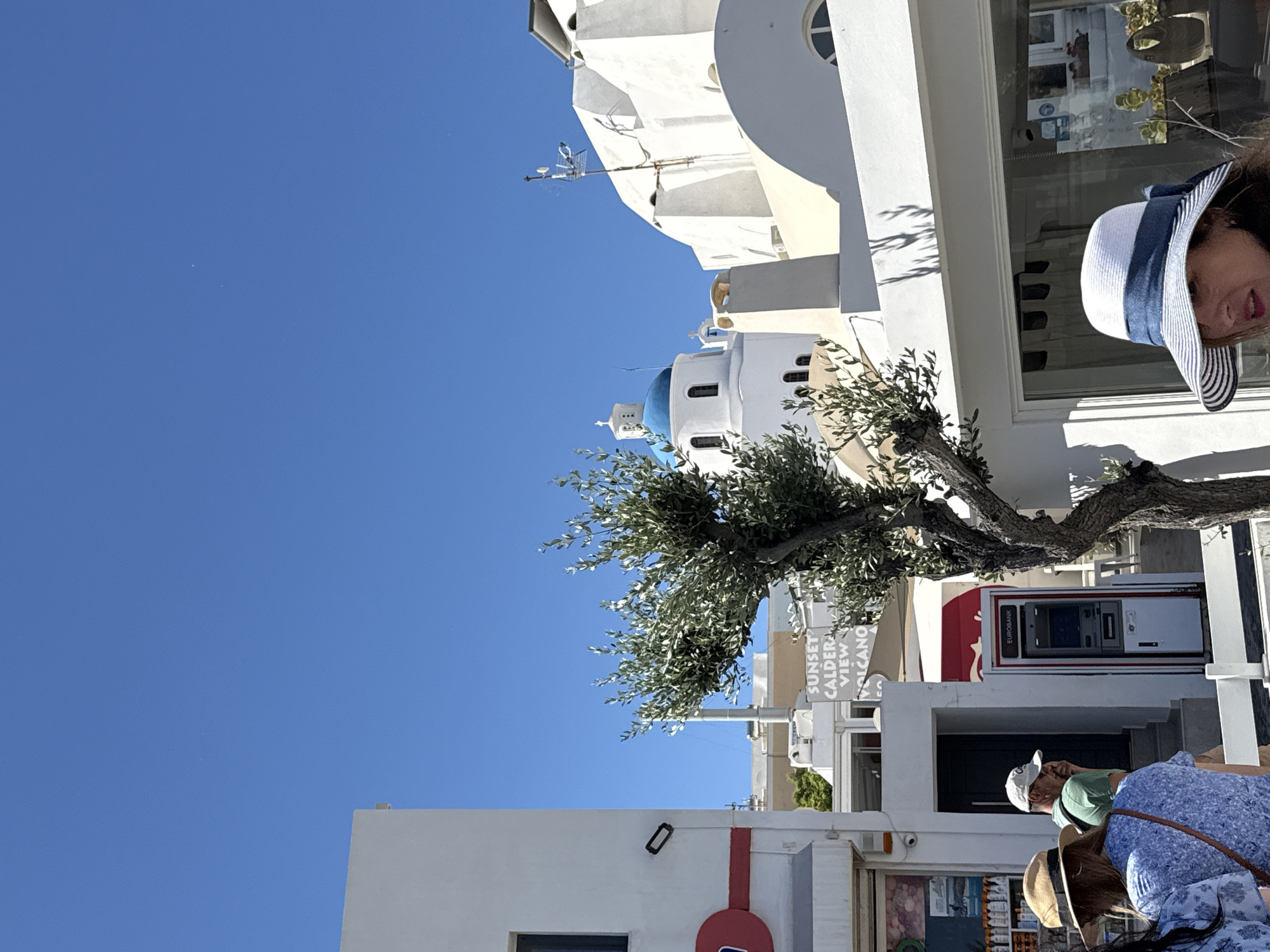 In Ia Municipal Unit, Greece, on a bright, sunny day, a scene unfolds on a street. A white building with a red stripe runs along the left side of the frame. To the right of this building, an olive tree with a gnarled trunk stands prominently. Beyond the tree, white buildings characteristic of Greek island architecture dominate the background, featuring domed roofs and arched windows. A prominent blue-domed church with a white cross stands out against the clear blue sky. In the foreground, several people are visible. A woman wearing a straw hat with a blue ribbon is partially in the frame on the right. To her left, a woman in a blue floral top and a straw hat is seen from behind. Further left, a man wearing a green shirt and a white baseball cap is talking on his phone, looking towards a building entrance with stairs. An ATM machine, branded "EUROBANK," is located to his right. Signs for "SUNSET CALDERA VIEW VOLCANO" are visible behind the olive tree, indicating the area's attractions. The overall impression is a bustling street scene in a picturesque Greek town.