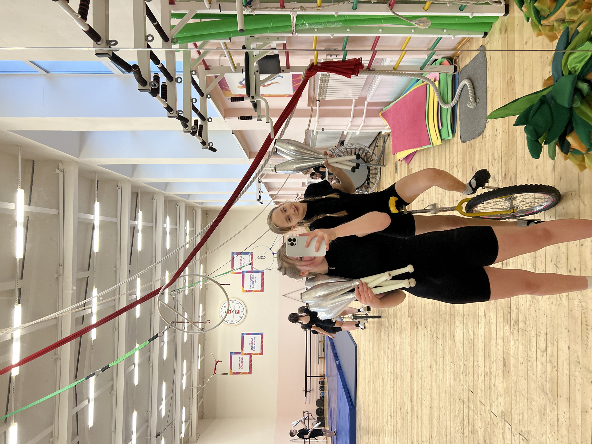 The image shows an indoor gymnasium-like space in Chaikovsky, Russia. Two young women are riding unicycles and juggling clubs, likely practicing or performing. One woman is holding a smartphone, presumably taking a selfie of themselves with the other juggler. The gymnasium is equipped with gymnastic equipment and there are some fitness-related objects visible like weights and mats.