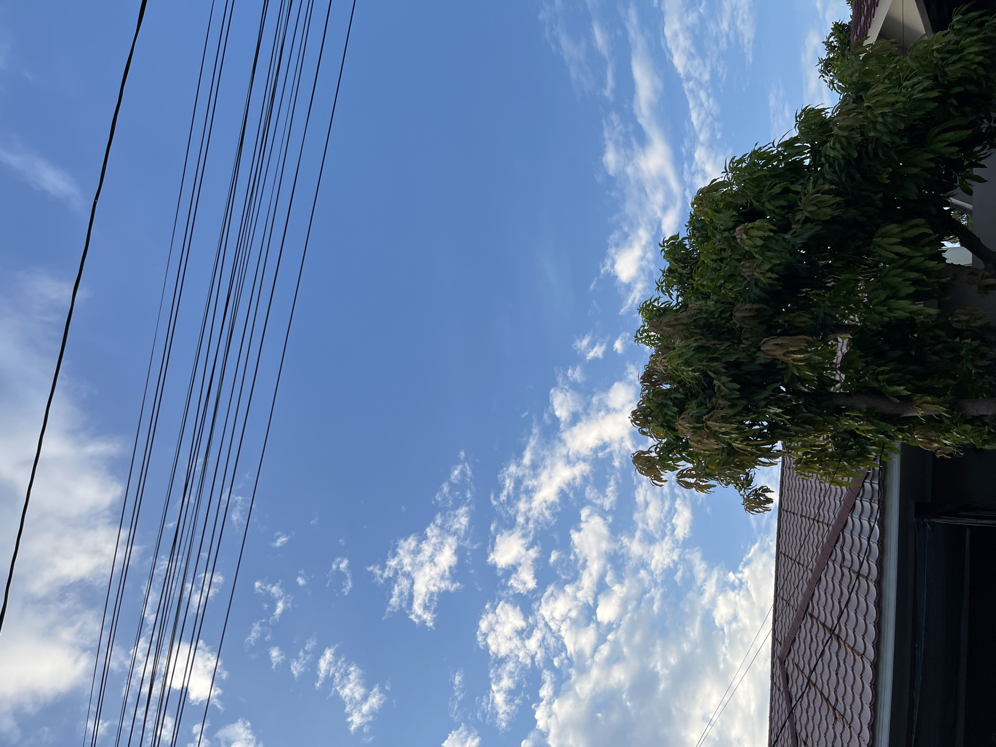 The image depicts a residential scene in Lhokseumawe, Indonesia, under a partly cloudy blue sky. In the foreground, a portion of a house with a brown tiled roof is visible, extending from the left to the right edge of the frame. To the right of the house, a large tree with lush green leaves dominates the lower right quadrant of the image. Above the house and tree, several parallel black wires stretch horizontally across the sky, suggesting the presence of utility lines. The sky itself is a vibrant blue, interspersed with scattered white clouds, some of which are wispy and thin, while others are more dense and puffy. The overall impression is a serene, daytime view of a suburban or residential area.