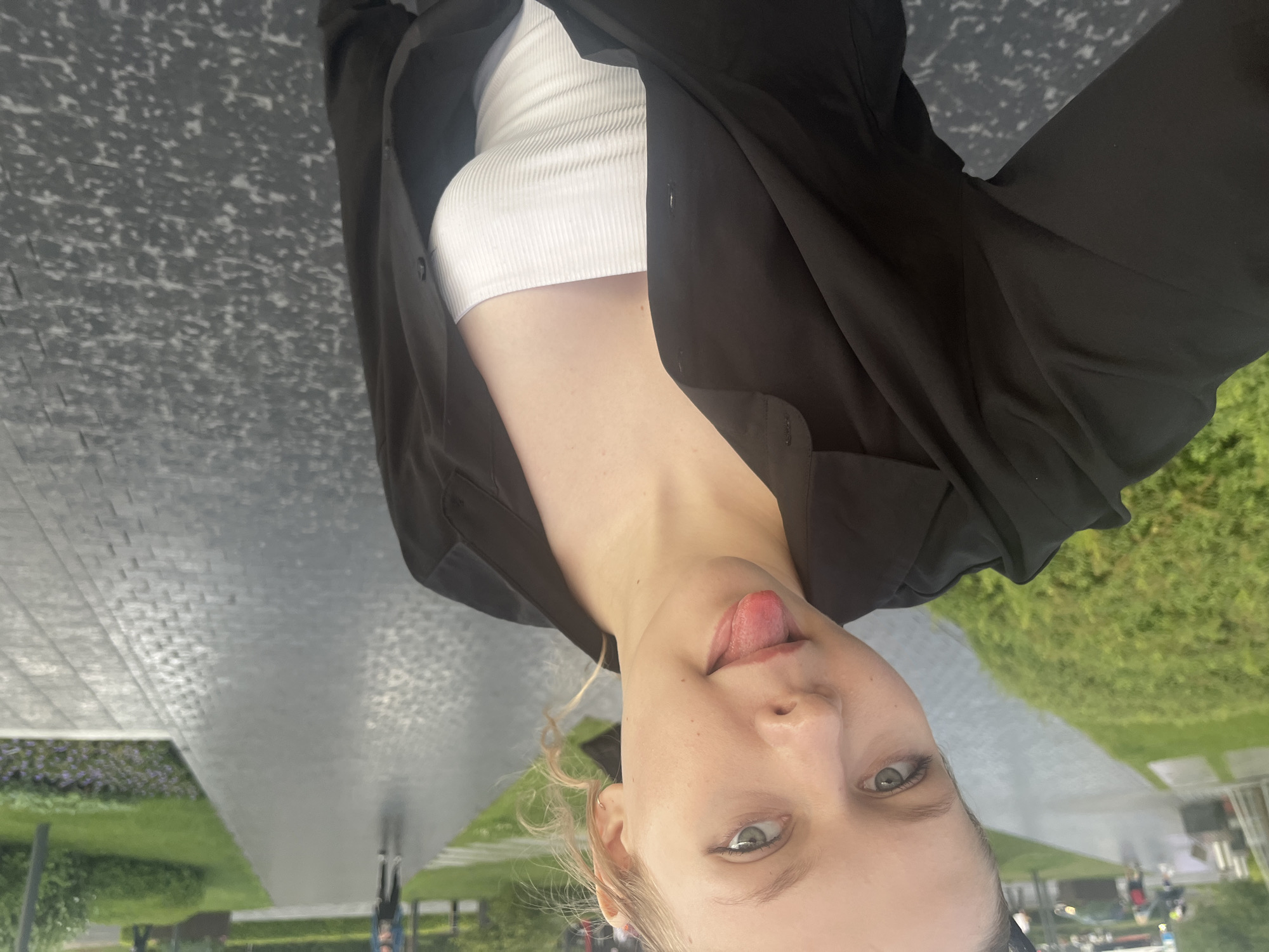 A young Caucasian woman with blonde hair tied back and her tongue sticking out is taking a selfie outdoors. She is wearing a black open button-up shirt over a white ribbed tank top. The background shows a paved walkway with cobblestone patterns, green grass and bushes, and some people walking in the distance. The overall setting appears to be a park or public walkway in the city of Perm, Russia.