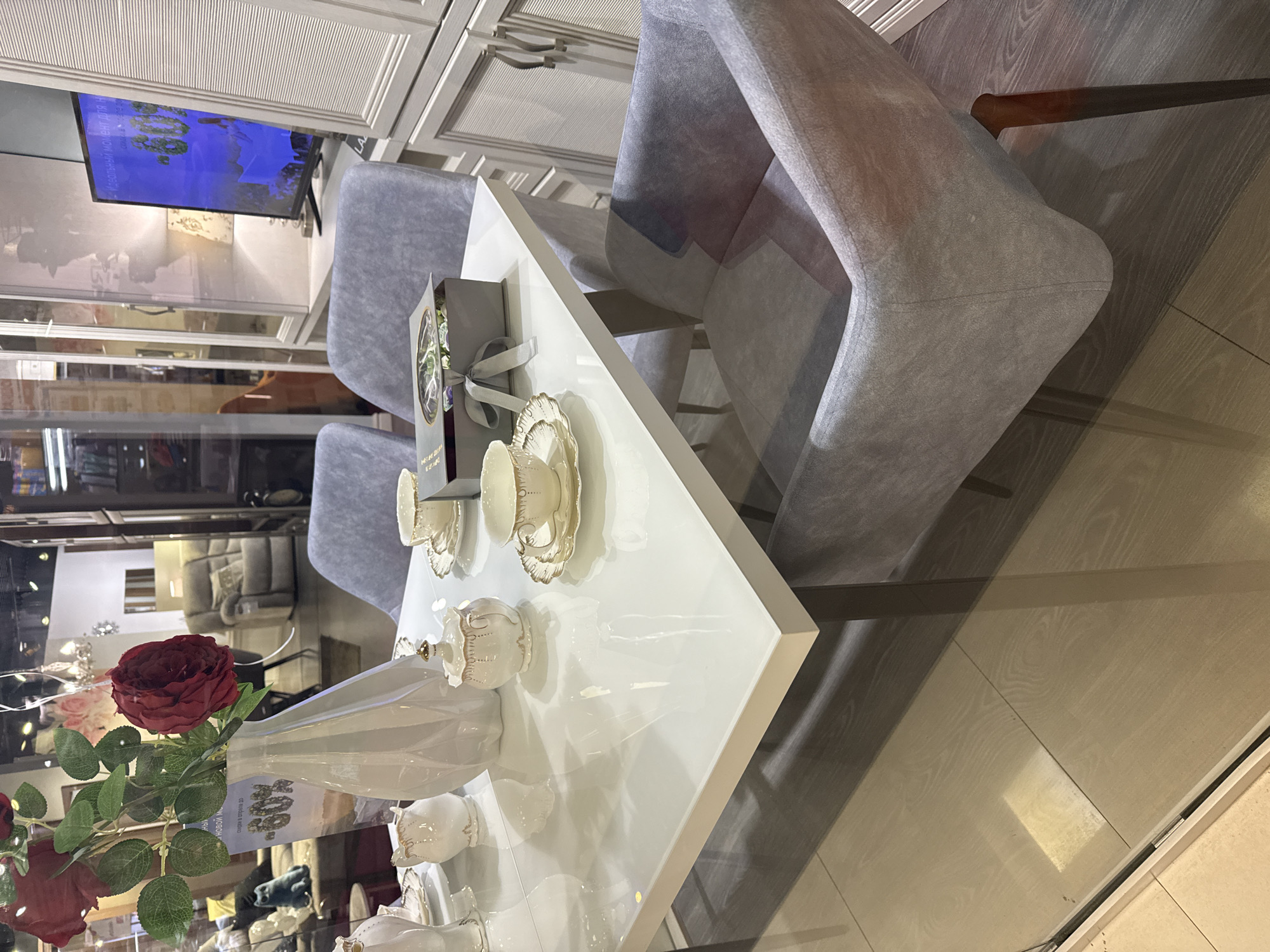 The image shows a display in Dmitrov, Russia, likely within a furniture or home decor store. A white, square table is set with fine porcelain teacups, saucers, and a creamer. A tall, white vase containing red roses is also on the table. Behind the table are two gray upholstered chairs. To the left, a television screen is visible, displaying a video. Shelves and other furniture are behind the chairs. The flooring appears to be wood laminate. The scene is well-lit, suggesting an indoor retail environment. A gift box with a ribbon is placed between the table and the chairs.