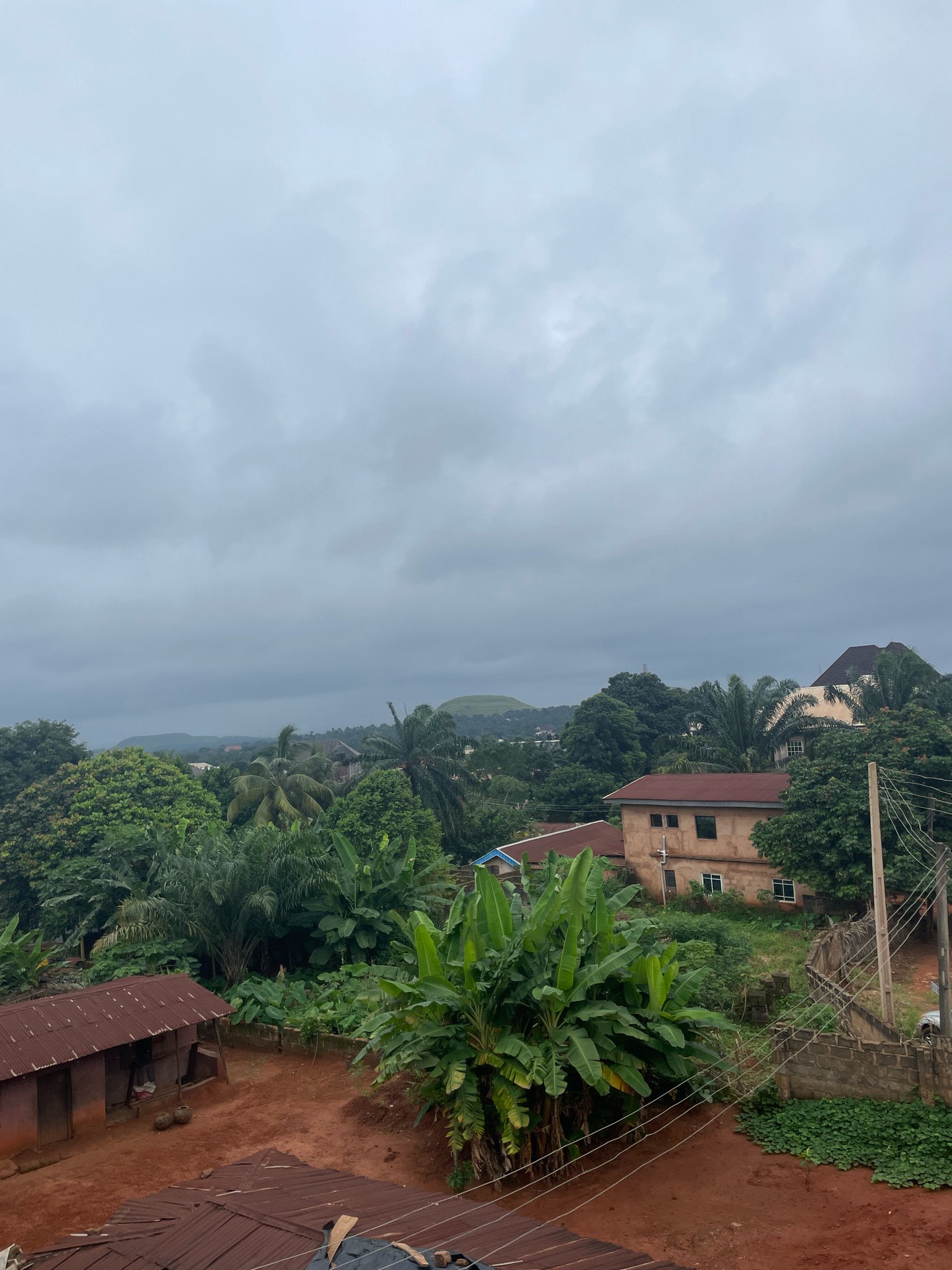 The image shows a landscape in the city of Nsukka, Nigeria, under a cloudy sky. In the foreground, there are two structures with corrugated metal roofs, one appearing to be a small, rustic dwelling. Behind these, a lush green environment dominates, with dense vegetation including banana trees and palm trees. Further in the background, several buildings are visible, some with reddish-brown roofs and beige walls. A hilly landscape with a distinct rounded peak is visible in the far distance, with more buildings interspersed among the trees. Power lines and a utility pole are present on the right side of the frame, suggesting some infrastructure in the area. The ground is mostly red earth, with some areas covered in greenery. The overall scene depicts a semi-urban or rural setting within Nsukka, characterized by abundant vegetation and a mix of traditional and modern housing.