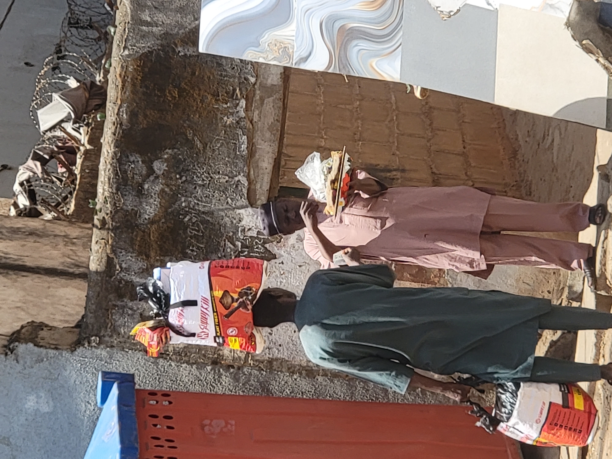 This outdoor scene in Maiburiji, Nigeria, captures two men engaged in daily activities, likely transporting goods under bright sunlight.
The primary subjects are two adult males, both appearing to be carrying items on their heads. The man on the left, dressed in dark green traditional attire (a long tunic and trousers), carries a large, white and red sack prominently on his head. Visible text on this sack includes "SUNNY KILL," "PLANT FIBER," and "GOOD QUALITY," accompanied by graphics that suggest agricultural products like seeds or grains, and a tool resembling a hoe. The man on the right, wearing a light brown or pink long-sleeved shirt and trousers, along with a dark cap, also balances a smaller, lighter bundle on his head. This bundle appears to be wrapped in clear plastic, with thin sticks or other items protruding, and he is holding something small to his mouth or nose.
The activity depicted is the act of carrying goods, a common sight in local markets or street environments. The men seem focused on their task, possibly en route to a market or a delivery.
Notable details include the bright, clear daytime lighting, indicated by the distinct shadows cast on the ground and walls. The background features a textured wall, possibly concrete or rough stone, suggesting an older building or construction. Above the wall, various patterned tiles are visible, which could be part of a building's facade or flooring displays. In the foreground, parts of an orange and blue structure, possibly a barrier or utility box, are visible. The overall setting suggests a bustling, functional urban or semi-urban environment.