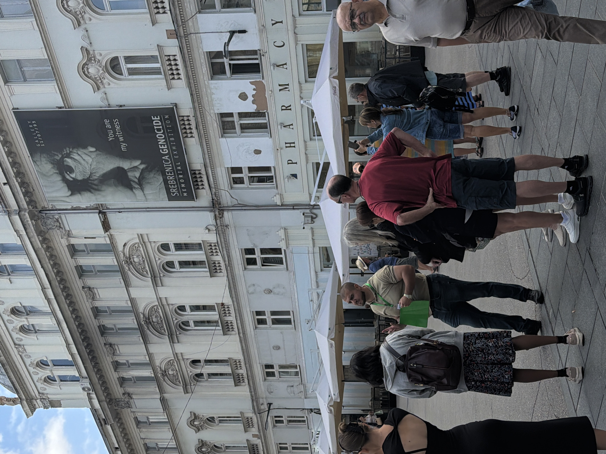 The image depicts a street scene in Sarajevo, Bosnia and Herzegovina, focused on a building facade displaying a large banner for the "Srebrenica Genocide Memorial Exhibition." Several people are visible in the foreground, standing on a paved area.
The building is multi-story with ornate architectural details. A prominent banner on the upper portion of the building features a black and white close-up of an eye and text that reads "You are my witness" and "SREBRENICA GENOCIDE MEMORIAL EXHIBITION." The date "GALLERY 11/07/95" is also present. Below the banner, the word "PHARMACY" is visible in large letters above a street-level entrance. White umbrellas, likely from outdoor seating for a cafe or restaurant, are positioned in front of the building.
In the foreground, a group of individuals are gathered. On the far left, a woman in a black dress is partially visible. Next to her, a woman with her back to the camera wears a grey hoodie and a patterned skirt, with a backpack. A man in a tan t-shirt and jeans is speaking, holding a green bag. To his right, a woman with blonde hair in a denim jacket is looking at her phone. Further to the right, a man in a red t-shirt and shorts stands with his hands on his hips. Another man in a dark jacket and shorts is also present, as is a man in a light-colored polo shirt and brown pants on the far right. The ground is paved with square tiles. The overall lighting suggests an overcast or partly cloudy day.