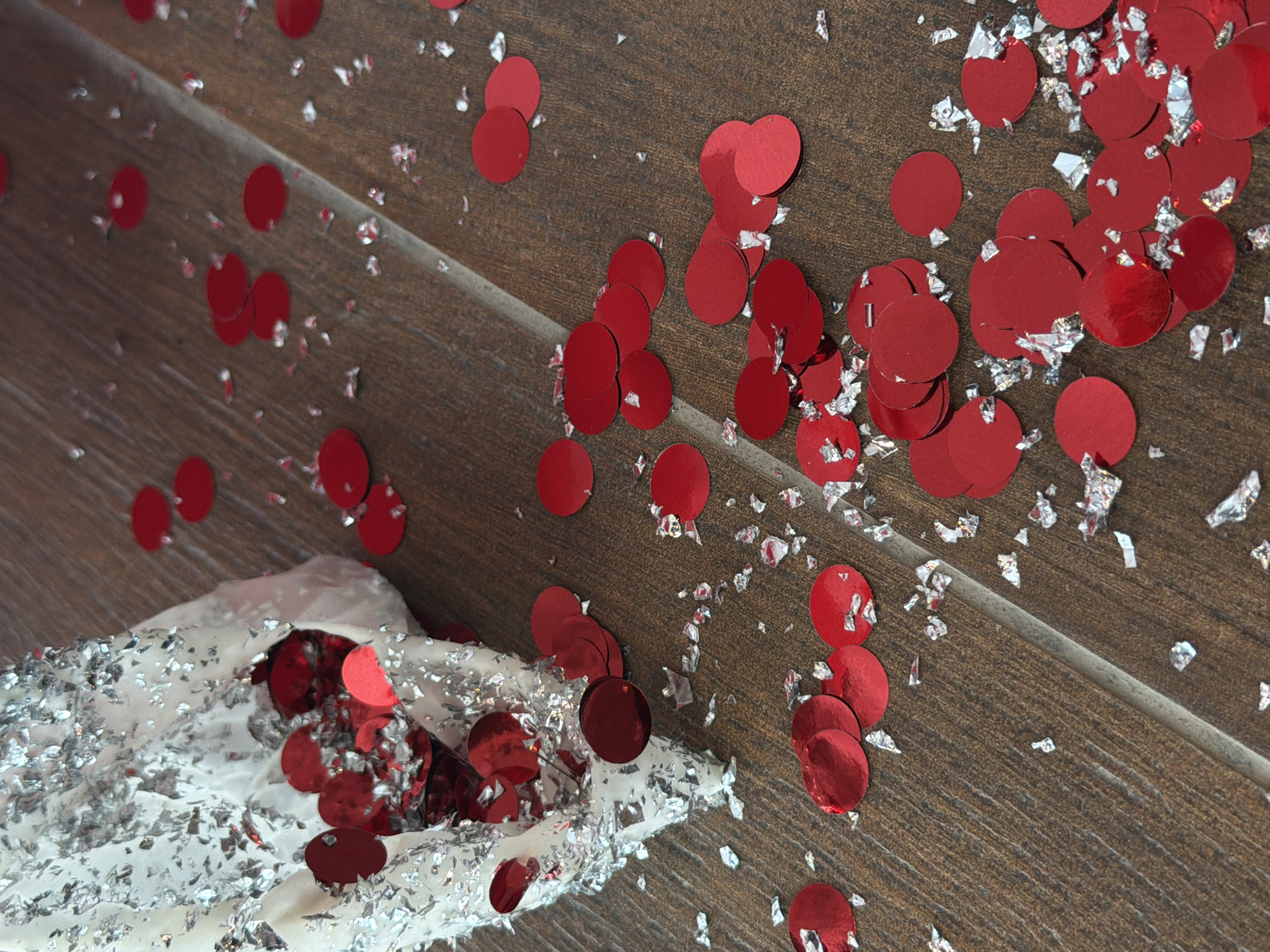 The image depicts a wooden surface covered with red circular confetti and silver tinsel. The confetti and tinsel are scattered across the surface, with a concentration near what appears to be a partially obscured object wrapped in white material and tinsel. The wood grain is visible, and the overall impression is one of celebration or post-celebratory mess. The location is not discernible from the image.