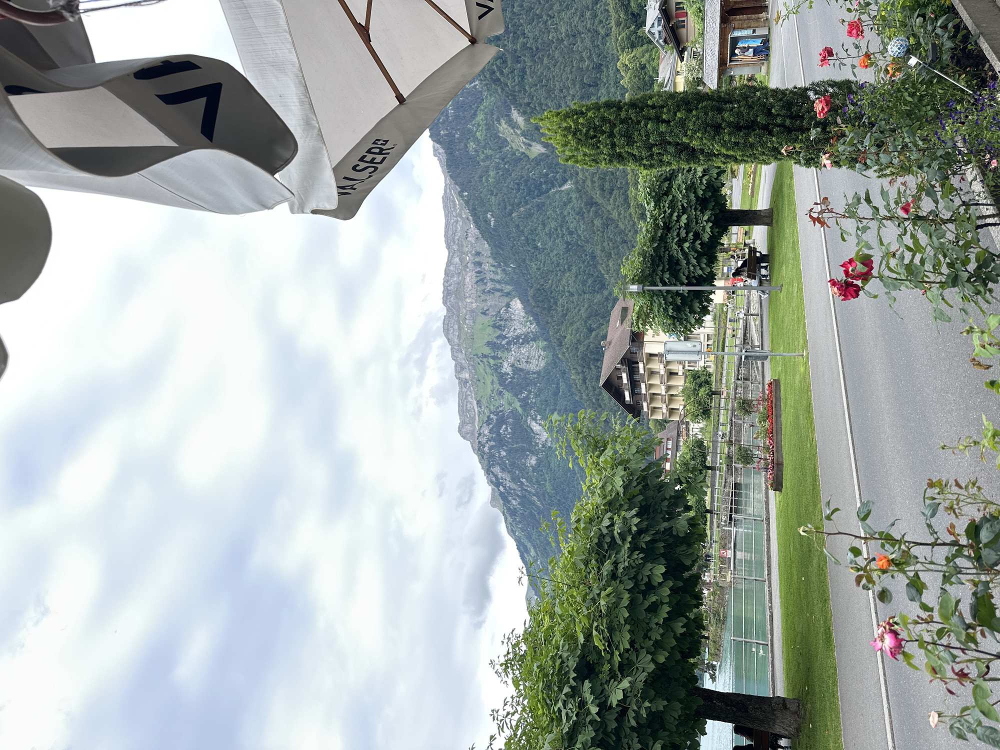 This image captures a view from a covered seating area looking out towards Lake Brienz in Bönigen, Switzerland. A white umbrella, with the "Walser" logo visible, shades the foreground. Beyond the umbrella, a road runs parallel to the lake, with a grassy embankment and a walkway. Trees line the walkway, and in the distance, a multi-story building with balconies is visible. The lake is a vibrant turquoise color, reflecting the cloudy sky. Lush green mountains form the backdrop, their slopes covered in dense forest. In the immediate foreground, several rose bushes are in bloom with red and pink flowers. A few people are seated on a bench near the lake, and others are walking along the path. The scene is serene and picturesque, typical of a lakeside resort town.
