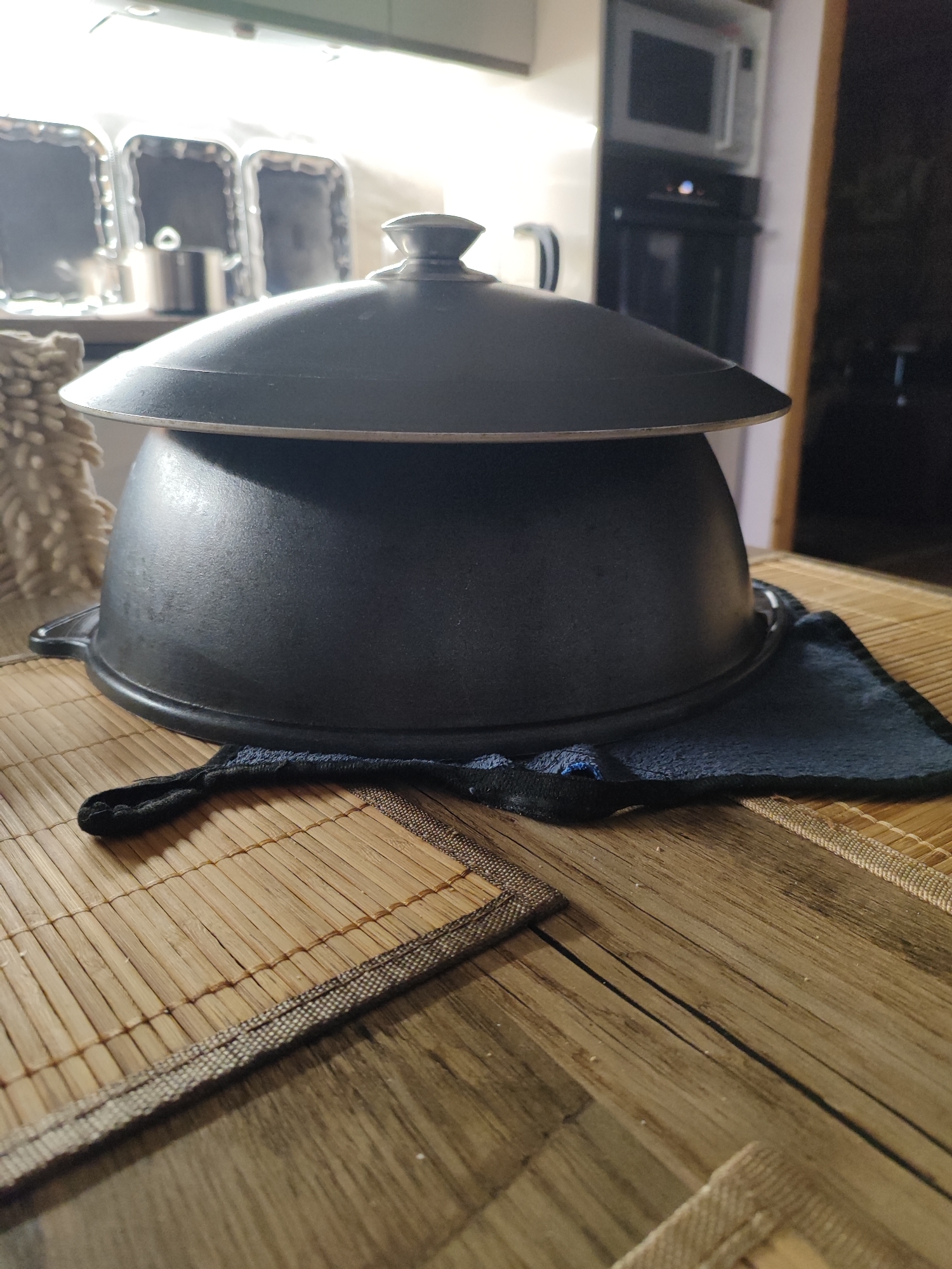 A black pot with a lid is placed on a bamboo mat on a wooden table. The pot appears to be made of cast iron. A dark cloth is underneath the pot, on top of the bamboo mat. In the background, a microwave and some kitchen cabinets are visible. The scene is indoors, likely in a kitchen. The image does not provide information about the city of Marusino, Russia.
