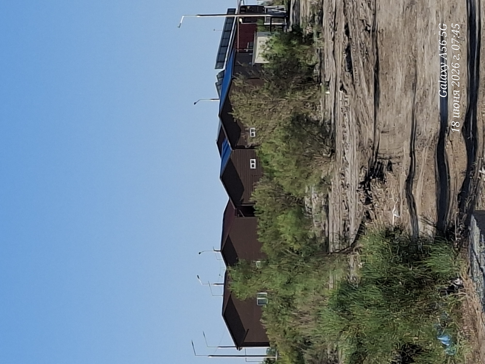 A medium shot captures a cluster of modern, dark brown, wooden houses with dark blue solar panels on their roofs under a clear blue sky. The houses are nestled among dry, scrubby vegetation and dusty, uneven ground, suggesting a semi-arid or developing environment. Several tall, modern streetlights stand around the buildings. The foreground shows a rough, dirt track. In the lower right corner, text indicates "Galaxy A56 5G 18 июня 2026 г. 07:45". The scene appears to be a residential area in a sparsely vegetated, possibly rural or suburban location in Uzbekistan.