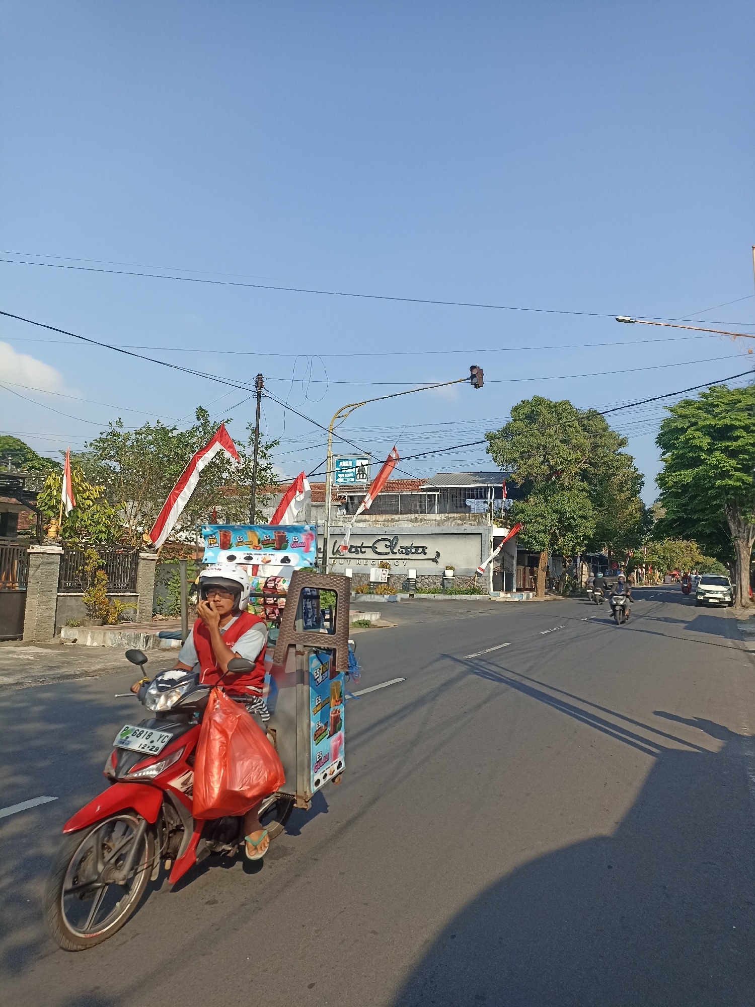 A person rides a red motorcycle with a food cart attached. The motorcycle is on a paved road in Mojopanggung, Indonesia. The person is wearing a red shirt and a white helmet. The food cart has an advertisement for "Es Teh" and "Es Kul" ice drinks. In the background, there are trees, buildings, and Indonesian flags. The sky is clear and blue.