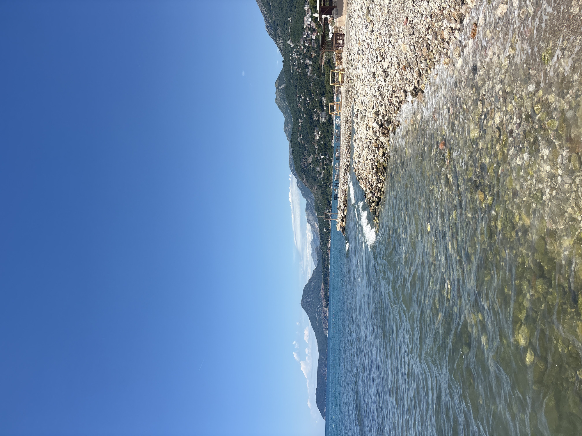 The image depicts a coastal scene in Bar, Montenegro, under a clear blue sky. The foreground features a pebble beach with clear, shallow water lapping at its edge, revealing submerged stones. The water is calm with gentle waves. Beyond the beach, a rocky breakwater extends into the sea, with a few beach chairs and umbrellas visible further down the coast, suggesting a developed recreational area. In the mid-ground and background, verdant, tree-covered hills rise steeply, dotted with houses and buildings, characteristic of the Mediterranean landscape. The overall impression is one of a serene, sunny day at a seaside resort.