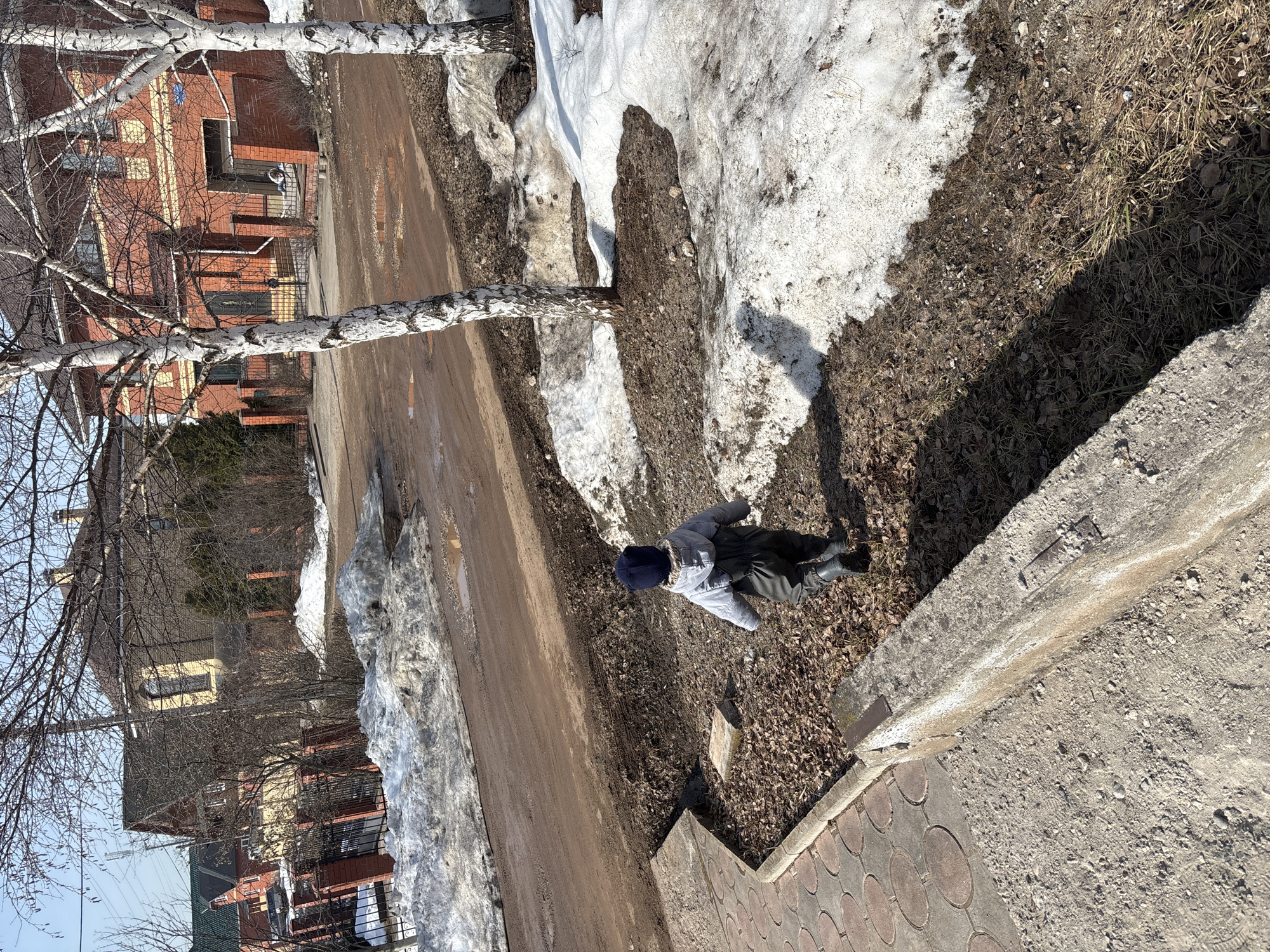 A medium shot depicts a person lying prone on a patch of dead grass and leaves adjacent to a melting snowbank along a street in Ферма, Russia. The individual wears a light gray jacket, dark pants, and a navy blue hat. The street is wet and muddy. Several buildings are visible across the street. A birch tree stands near the snowbank. The image is taken from an elevated position near a sidewalk.