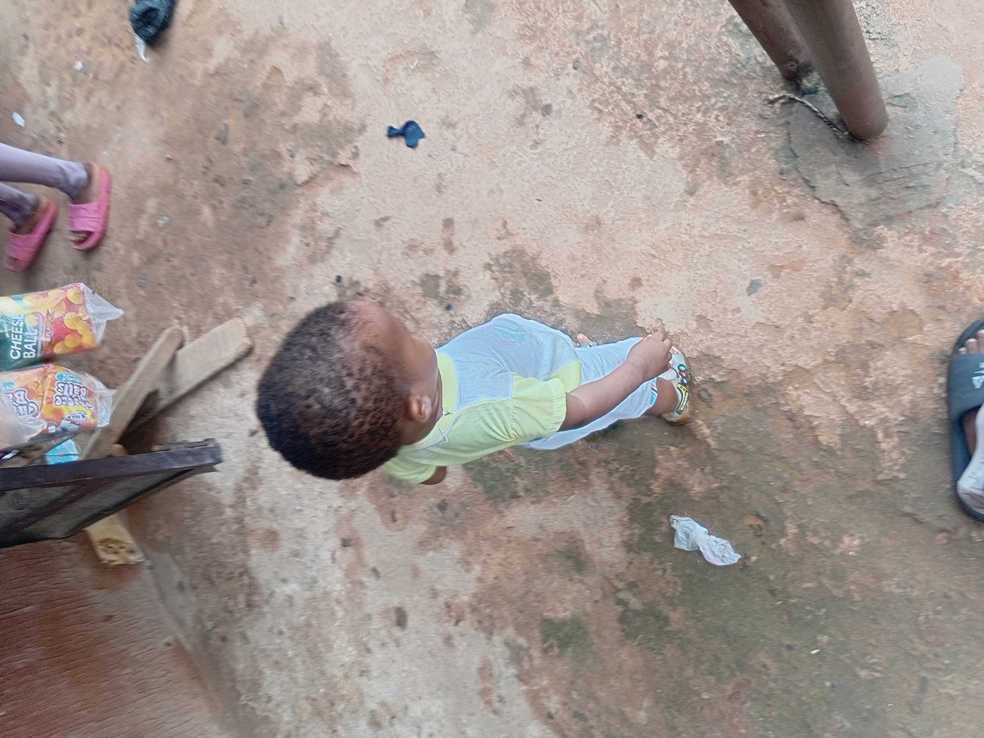 A child wearing a light yellow short-sleeved shirt and white pants is standing on a concrete surface. The child is facing away from the camera, looking down and to the right. The child's hair is dark and curly. Pink slides are visible on an adult's feet to the right of the child. A plastic bag with the words "CHEESE BALL" printed on it is to the left of the adult's feet. The scene appears to be outdoors in an urban environment, possibly Nkpor, Nigeria, with rough concrete ground and wooden structures in the background. A black plastic bag is on the ground to the far right.