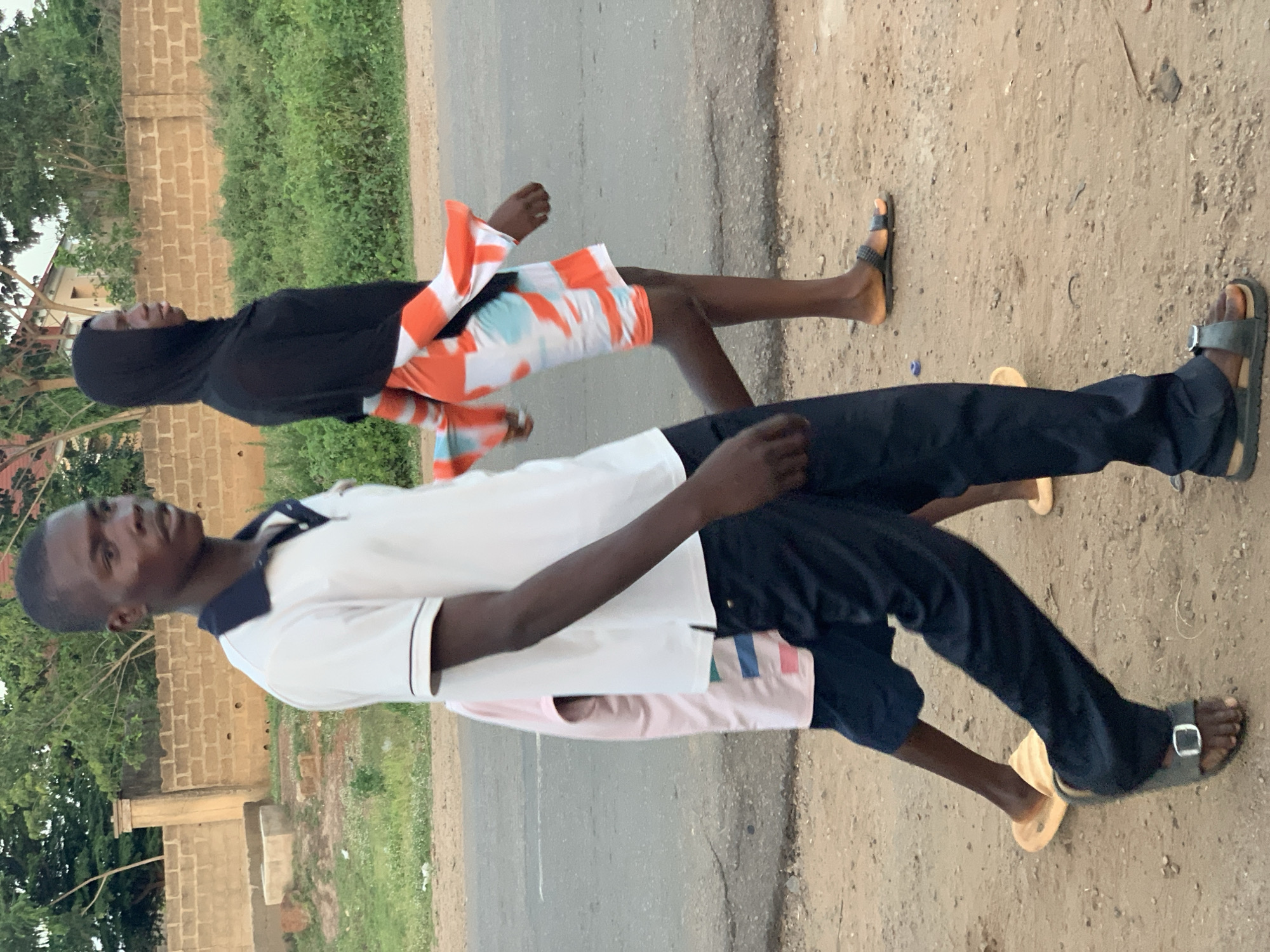 Two people are walking side-by-side on a paved road adjacent to a dirt shoulder in Kabba, Nigeria. The person in the foreground is wearing a white polo shirt with navy blue trim, dark blue pants, and open-toed sandals. The person in the background is wearing a black head covering and a short-sleeved top and shorts with an orange and white abstract pattern, and open-toed sandals. A low brick wall and green vegetation line the background.