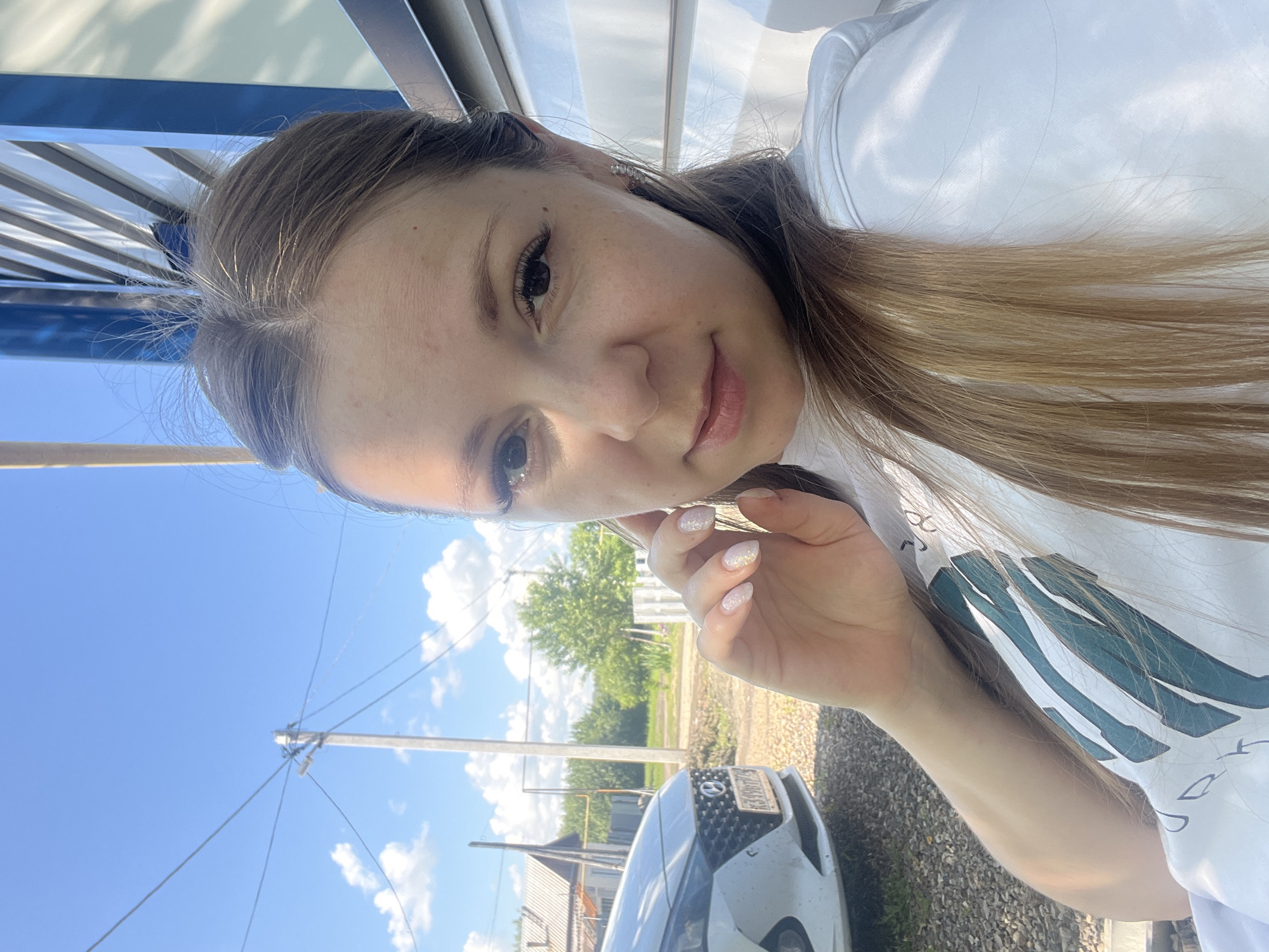 A young woman with blonde hair, light skin, and freckles poses for a selfie outdoors. She is wearing a white t-shirt with a green and white design. Her right hand is raised to her face, showing off sparkly nail polish. In the background, a white car is parked on a gravel surface. A wooden utility pole with electrical wires stands to the left, and a house with a brown roof and a white fence are visible further back. The scene is set against a bright blue sky with scattered clouds. The architecture and vehicles suggest a residential area in a rural or suburban setting, consistent with the city of Shadrinsk, Russia.