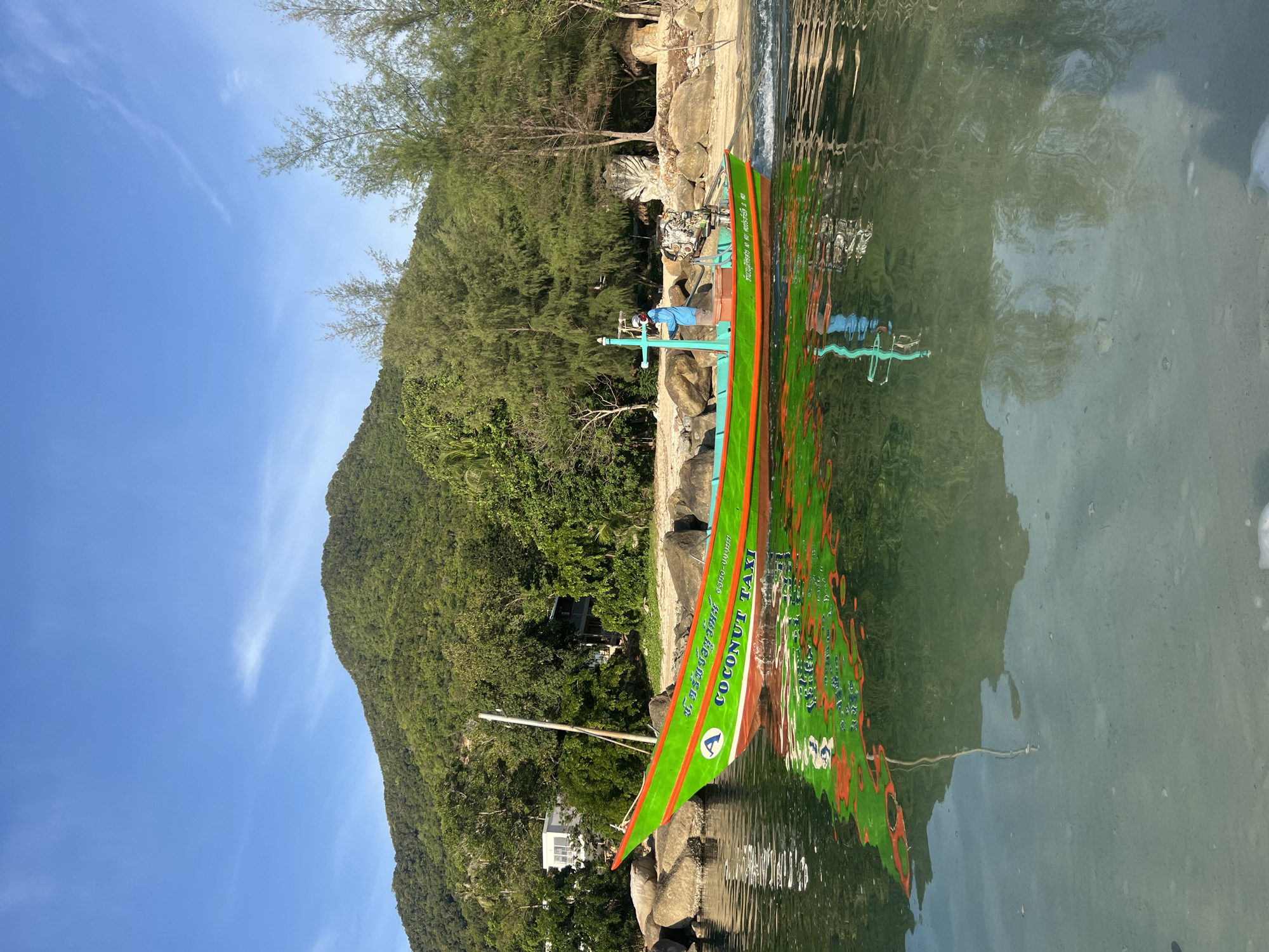 A bright green, long-tail boat named "COCONUT TAXI" with an orange trim is partially submerged in clear, calm water, reflecting the boat and the surrounding lush green mountainous landscape under a bright blue sky with wispy white clouds. The boat, which appears to be a fishing boat, has a man wearing a blue shirt and a light-colored hat standing near the back, operating the engine. The shore behind the boat is lined with large rocks and dense green trees. Some buildings are visible through the trees on the hillside. The water is shallow and transparent, revealing the sandy bottom. The scene is captured from a low angle, from the water's surface.