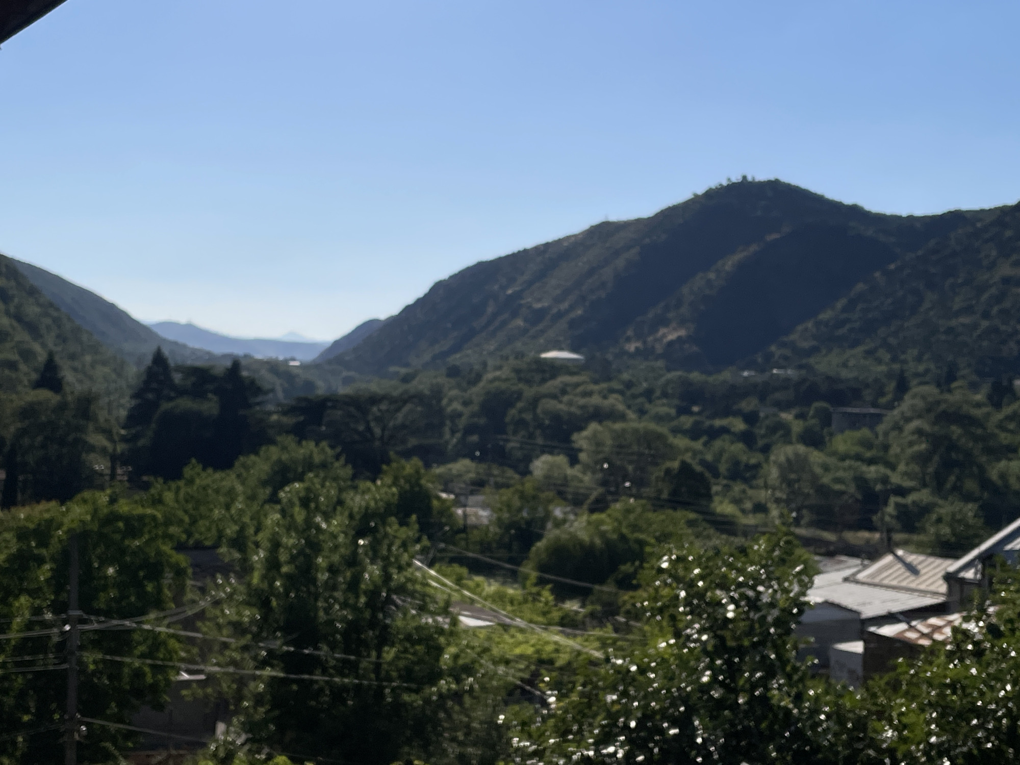 The image depicts a hazy, distant view of a lush, mountainous landscape with scattered structures, likely representing a settlement within or near the city of Mtskheta, Georgia. The foreground is dominated by the out-of-focus foliage of trees, obscuring a portion of the scene. In the mid-ground, a dense canopy of green trees creates a textured carpet that partially conceals buildings and what appear to be power lines. Several buildings with varied roof styles, some appearing aged with rusted corrugated metal, are visible amongst the trees. In the background, a series of progressively distant, shadowed mountains rise against a clear blue sky. One prominent mountain in the middle distance has a rounded, domed structure near its peak. The overall impression is one of a rural or semi-rural environment nestled within a mountainous region under a bright, possibly early morning or late afternoon, sky. The focus is soft, giving the image a dreamlike or impressionistic quality.