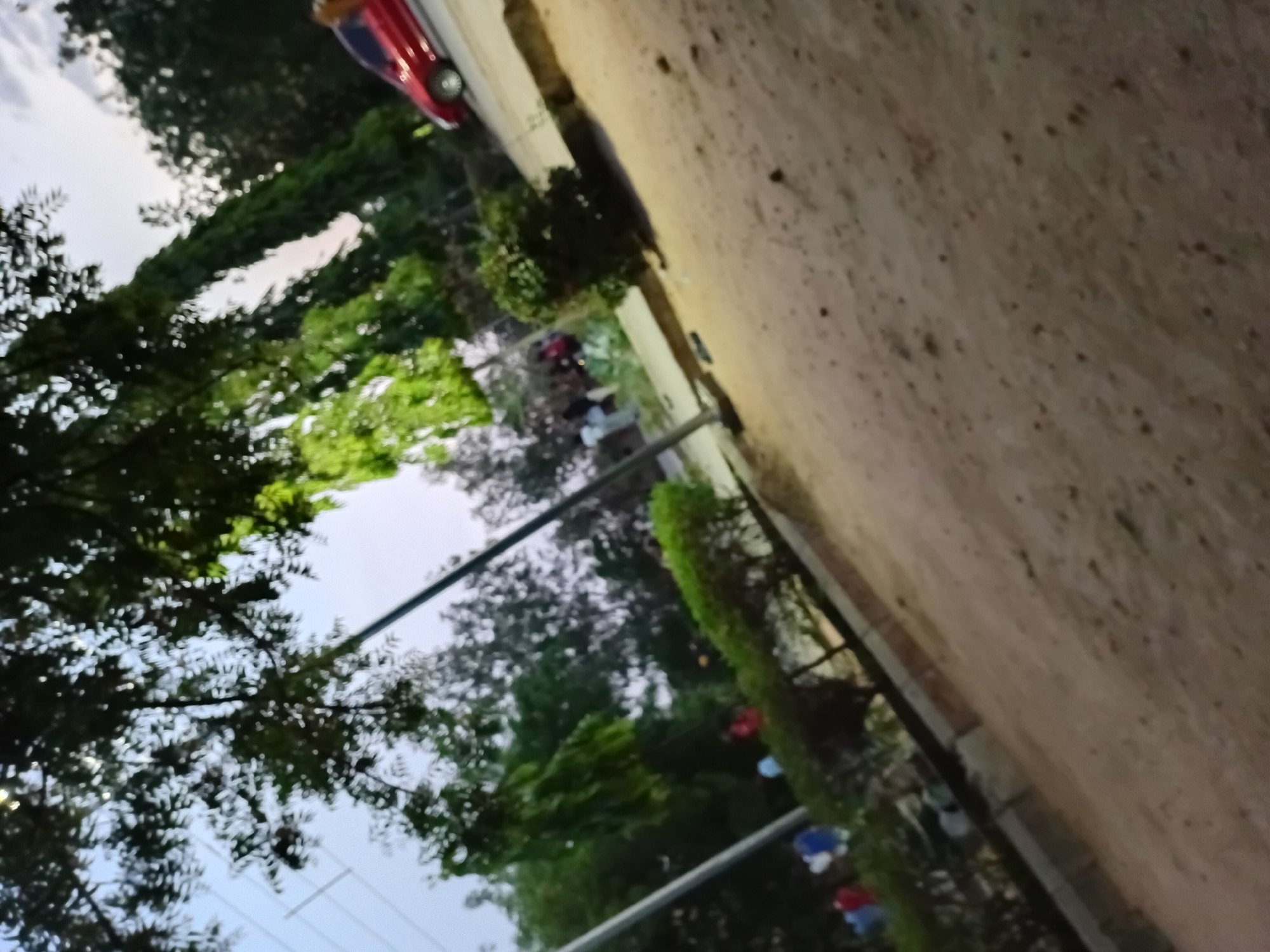 The image presents a landscape, seemingly taken outdoors. The scene includes various natural and man-made elements. There are trees with dense foliage visible, contrasting with the sky. A road, appearing unpaved, occupies a significant portion of the frame. The road surface seems to be composed of dirt or gravel. Along the road's edge, there is a boundary. There are individuals standing alongside the road, suggesting that this is a public area. The image includes a red car. The presence of overhead wires is indicative of urban infrastructure. The details of the surrounding city are difficult to discern given the available image data, but the combination of natural elements and infrastructural features suggests it is a residential or suburban area.