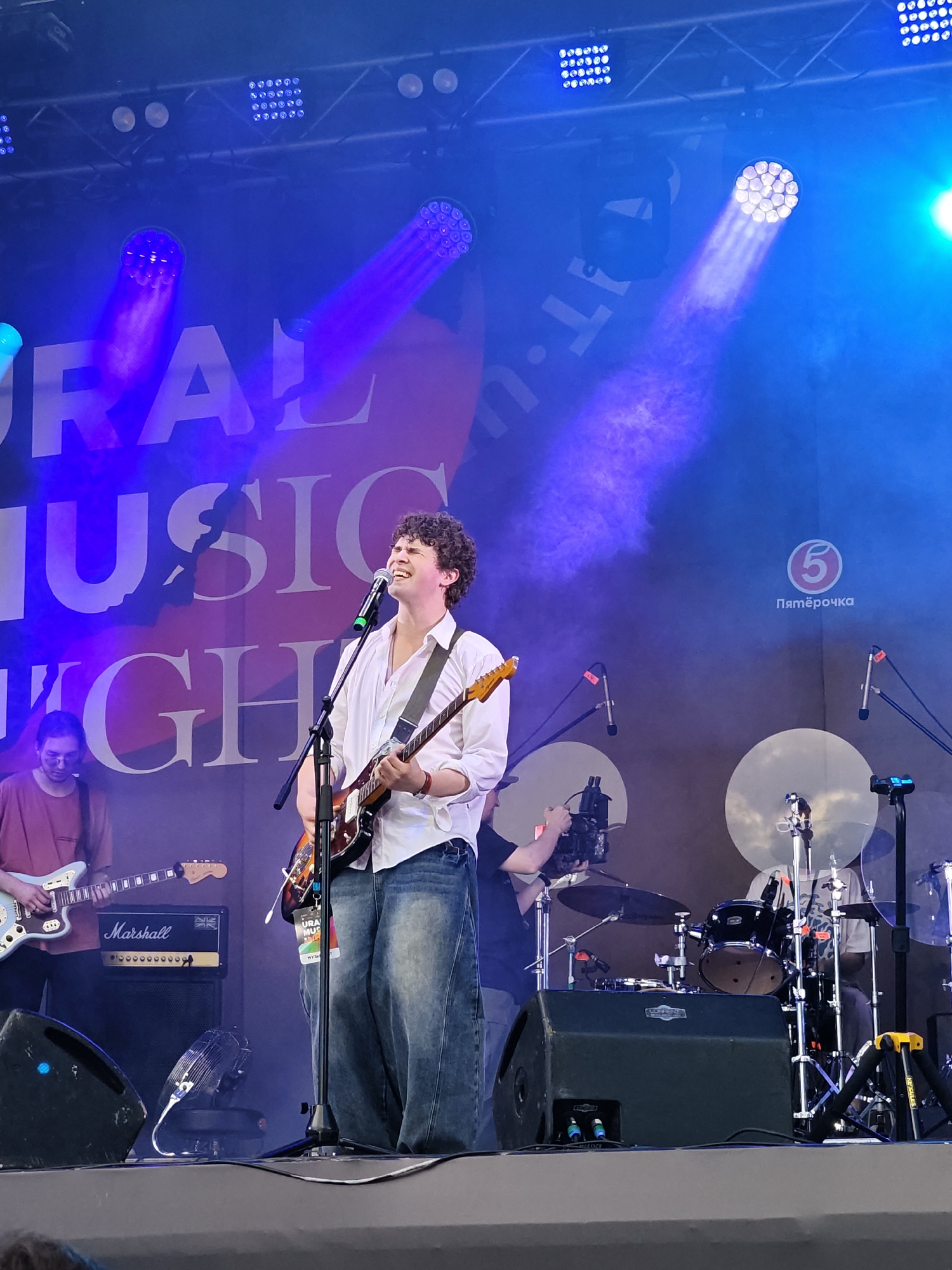 On a brightly lit outdoor stage, a young male musician with curly hair sings into a microphone while playing an electric guitar. He is wearing a white open-collared shirt and loose-fitting denim jeans. To his left, another musician plays an electric guitar, wearing a reddish-brown t-shirt. Behind them, a drummer is partially visible behind a drum kit. The stage is set with various lights, including spotlights that cast beams through smoke. A banner in the background features large white letters that read "URAL MUSIC NIGHT". The logo of "Pyaterochka," a Russian supermarket chain, is also visible on the right side of the banner. A Marshall amplifier is positioned on the stage. The scene appears to be from a music festival or concert in Yekaterinburg, Russia.