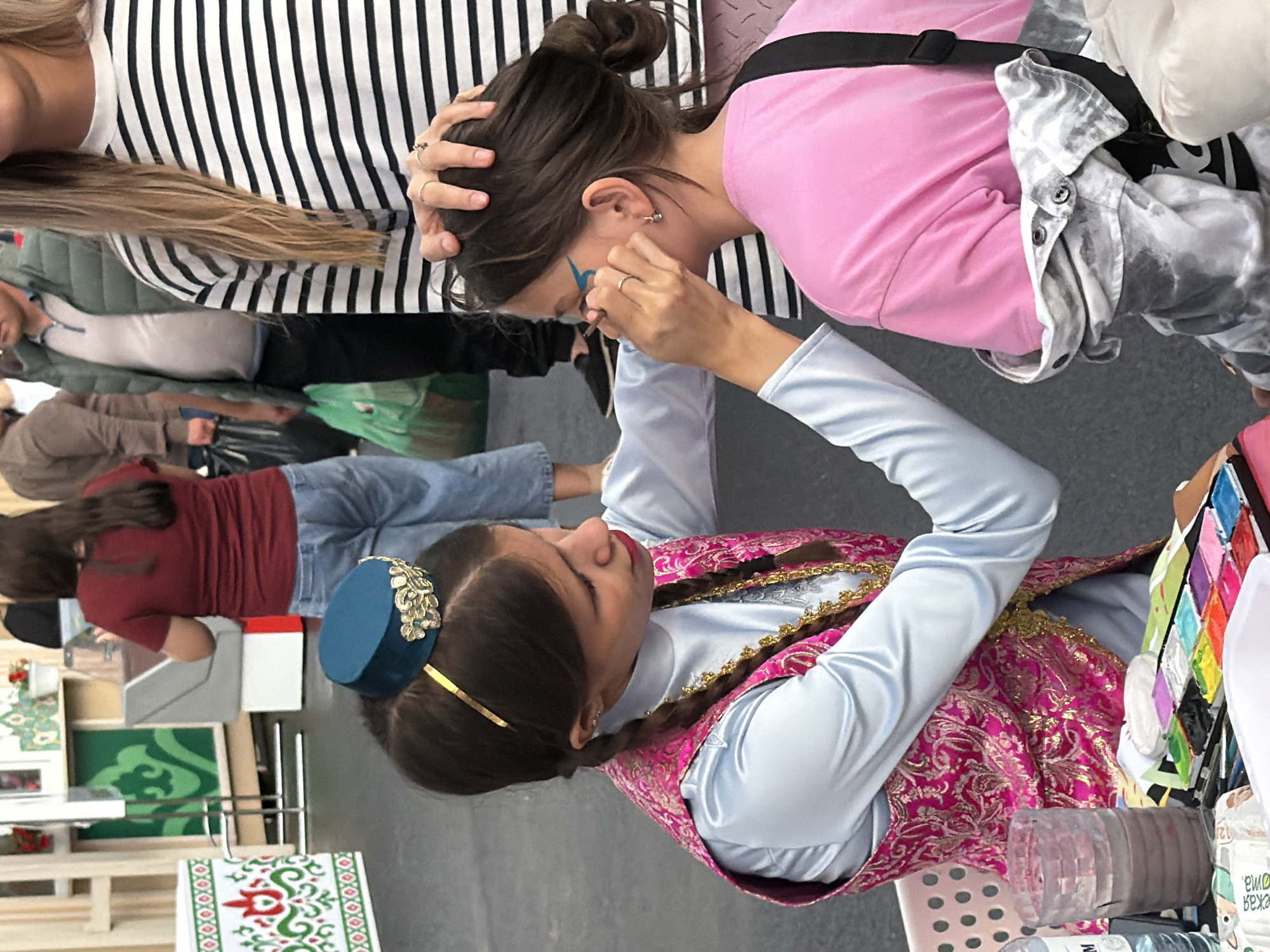 In an outdoor setting, a young person wearing a light blue shirt and a pink vest with gold trim is applying face paint to another young person's face. The face painter has dark hair styled in braids and wears a teal hat with gold embellishments. The person receiving the face paint has dark hair pulled back and is wearing a pink t-shirt and a tie-dye patterned jacket. The face painter is using a brush and a palette of colorful paints, with a cup of water nearby. Other people are visible in the background, some walking with shopping bags. The ground appears to be paved.