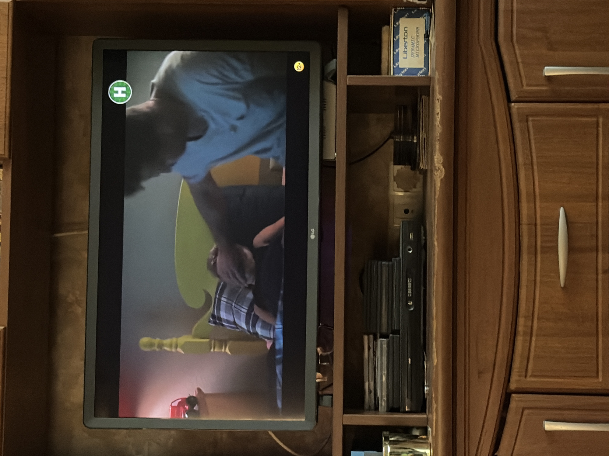 The image shows a television screen displaying a scene with two people. One person, wearing a light blue shirt, is bent over and appears to be holding a child. The child is wearing a plaid shirt and is visible from the chest down. A yellow chair is behind the child. A red object, possibly a toy, is on the floor near the bottom of the screen. A green circular logo with an "H" inside it is visible in the upper left corner of the screen, and a yellow circular logo with "12+" is in the upper right corner. The television is set into a wooden cabinet that also contains shelves with various items, including a box labeled "Liberton DYNAMIC MICROPHONE" and several stacks of what appear to be DVDs or CDs. The overall lighting suggests it is daytime, though the exact time of day is not specified. The setting appears to be a home entertainment area.
