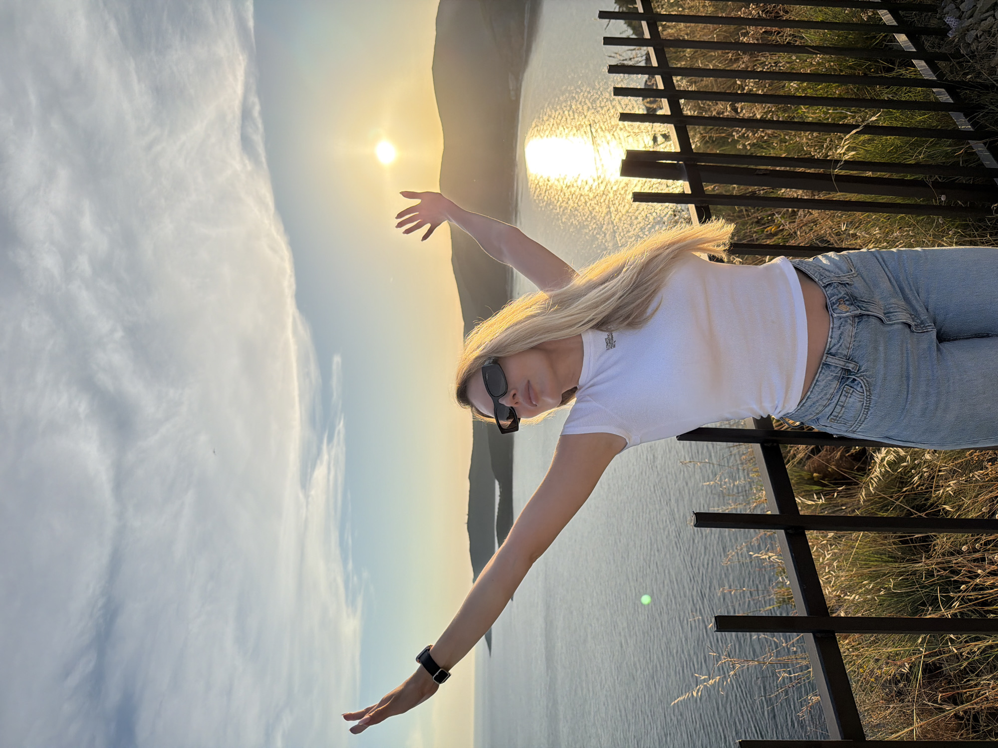 A woman with blonde hair is standing with her arms outstretched, facing the sunset over the Adriatic Sea. She is wearing a white t-shirt, blue jeans, and sunglasses. The scene appears to be on a hill overlooking the sea, with a black metal fence in the foreground. The sun is setting behind a mountainous coastline, casting a golden glow on the water. The location is identified as Budva, Montenegro, likely near the coast, perhaps on a viewpoint overlooking the bay.