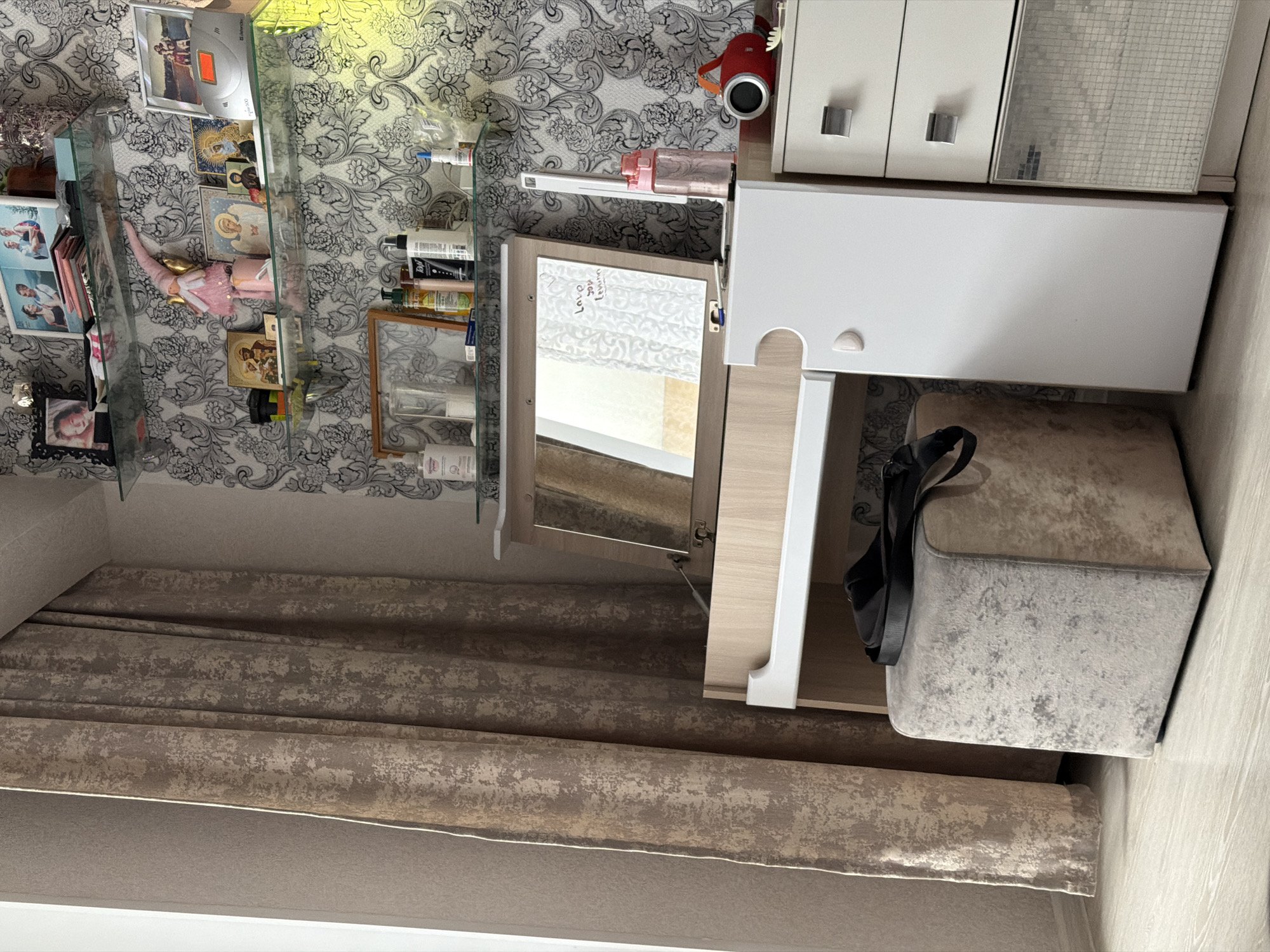 The scene depicts a section of a room with several notable features. A curtain with a patterned texture hangs partially in view on the left side of the frame. On the wall above the curtain, there is wallpaper with a grey and white floral pattern. A glass shelf unit displays various items, including framed pictures, figurines, and bottles.
Adjacent to the shelving unit is a vanity table. It features a mirror, storage compartments, and a white countertop. A grey upholstered seat is positioned under the vanity table, with a black bag resting on top. To the right of the vanity, a cabinet with drawers is visible.
The scene provides no information to verify a location in the city of Noviy, Russia.