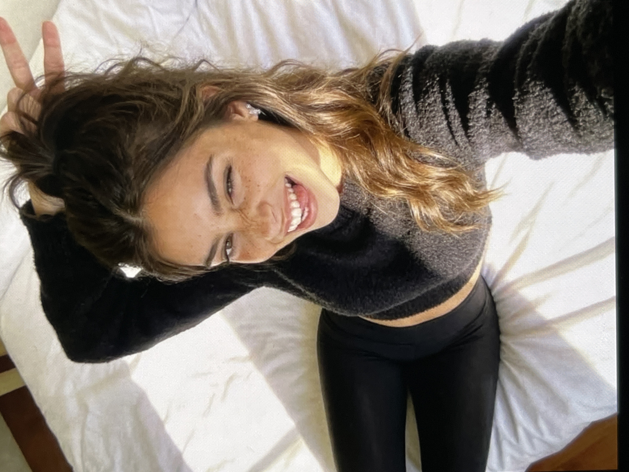 A young woman with long, wavy brown hair and freckles is lying on a white bed, looking up at the camera. She is wearing a black fuzzy sweater and black leggings. She is sticking her tongue out and winking one eye, while holding up two fingers in a peace sign with her other hand. The lighting suggests it is daytime, with sunlight casting shadows on the bed. There are no other people or objects in the immediate vicinity.