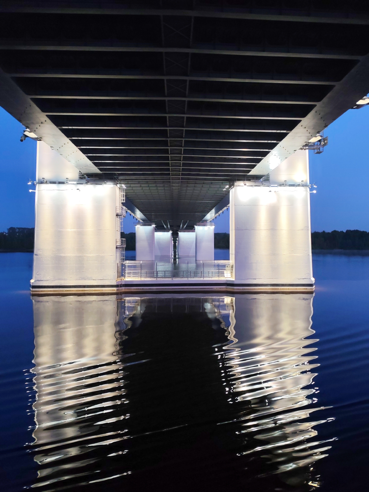 The image shows the undercarriage of a bridge illuminated at night. The supports are large, white, and rectangular, with lighting fixtures attached. Metal structures span horizontally beneath the roadway. The bridge is reflected in the dark water below, the reflections distorted by ripples. Trees are visible on the horizon in the background. The scene depicts a bridge at night in the city of Cherepovets, Russia.