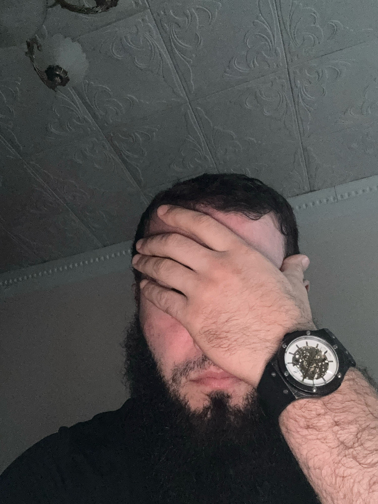 In a dimly lit room, a person with a beard covers their face with their right hand. A watch with a skeleton dial is visible on their left wrist. The ceiling above has a textured pattern and a light fixture. The image is set in Belidzhi, Russia.