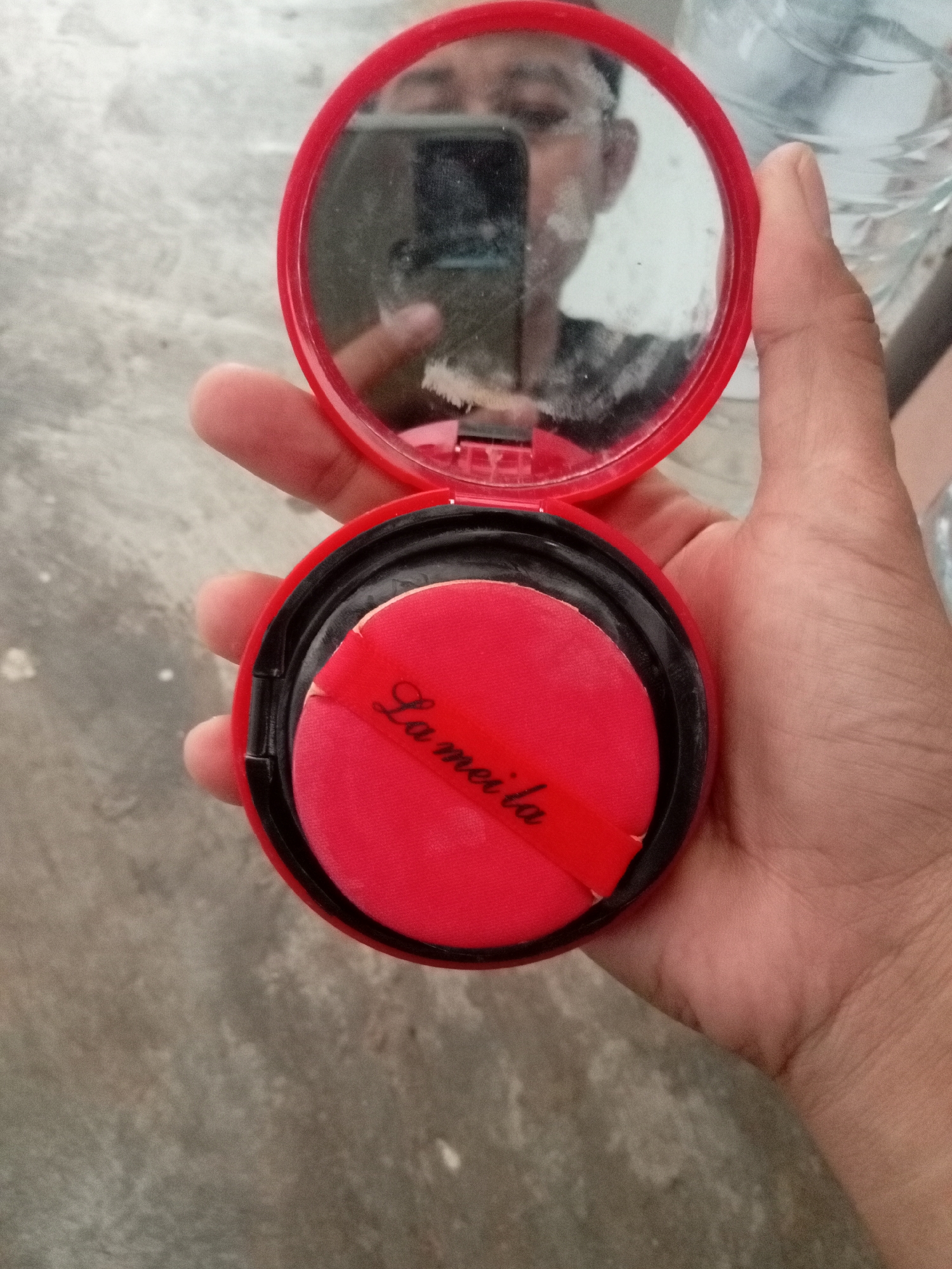 A person's hand is holding a compact powder case. The compact is red with a black inner rim. Inside, there is a red puff with the text "La meila" in black cursive script. The lid of the compact, which contains a mirror, is open and reflects a portion of the person's face and a cell phone. The background appears to be a concrete surface. The image is taken from a top-down perspective, and there are no discernible landmarks to indicate a specific location in Batam, Indonesia.