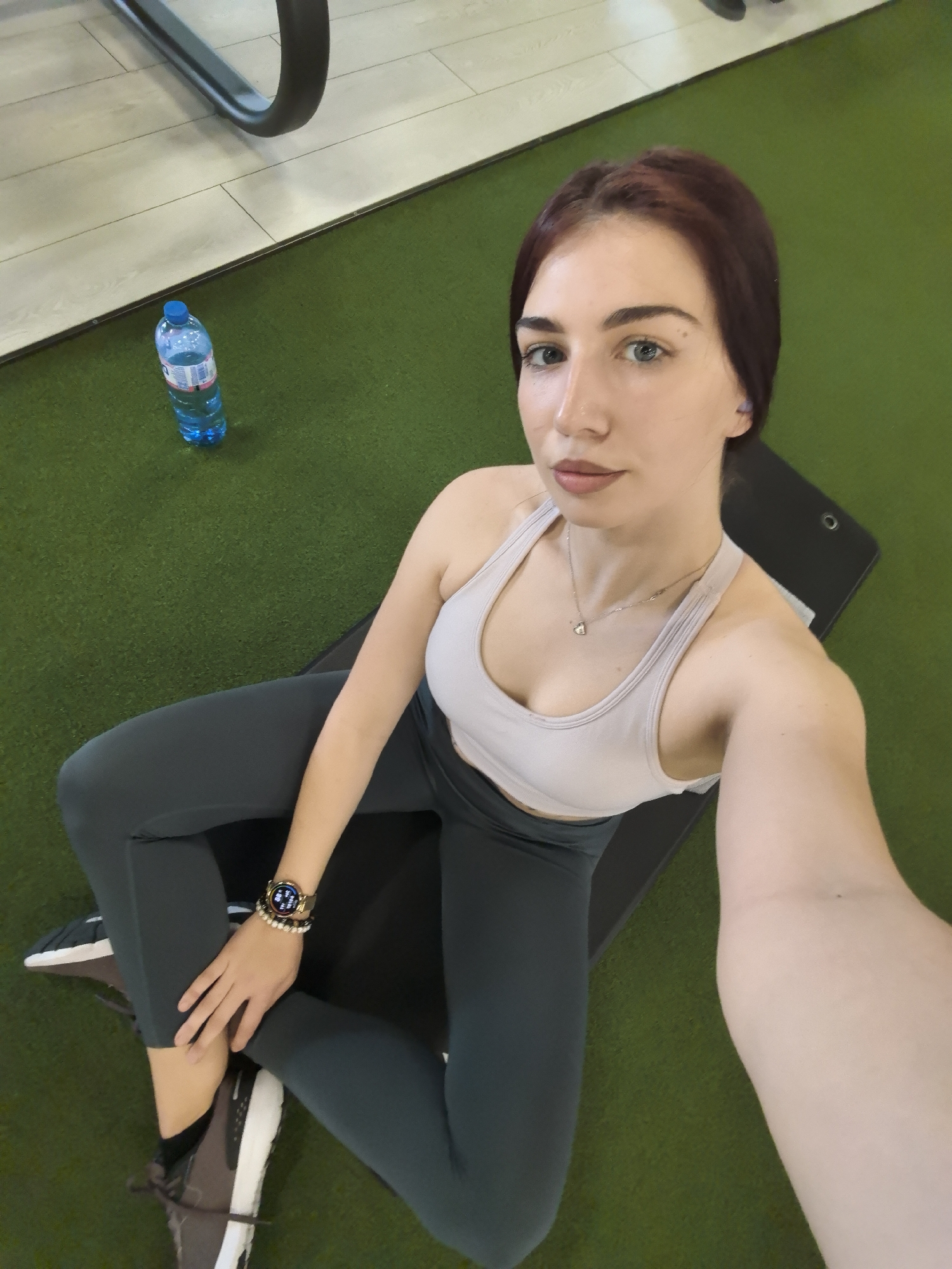 A young woman is sitting cross-legged on a black mat, positioned on green turf flooring. She is wearing a light-colored sports bra and dark leggings, with athletic shoes visible on her feet. A water bottle is located to the left of the mat on the green turf. The background also includes what appears to be a section of a gym with light-colored wood-look flooring. The location is Stara Zagora, Bulgaria.