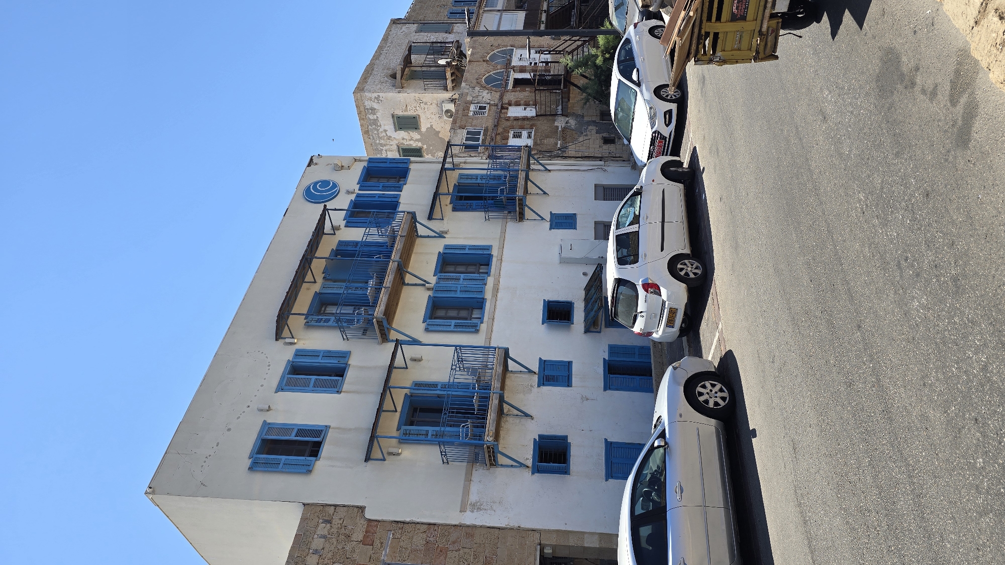 The image captures a street scene in the city of Acre, Israel, on a clear, sunny day. Dominating the left side of the frame is a white building with bright blue shutters on its windows and balconies. Several balconies, adorned with blue railings and some with outdoor seating, jut out from the building's facade. A distinctive blue circular logo with a wave-like design is visible near the top of this building. To the right, a slightly older, weathered building with similar architectural elements, including balconies and arched windows, stands adjacent. The street in front of the buildings is paved and appears to be a typical urban thoroughfare. Several cars are parked along the side of the street, including a silver sedan on the left, a white hatchback in the middle, and a white sedan on the right. A portion of a yellow utility vehicle with wooden planks loaded in its back is visible on the far right. The sky is a vibrant, cloudless blue. The overall impression is of a warm, Mediterranean coastal city.