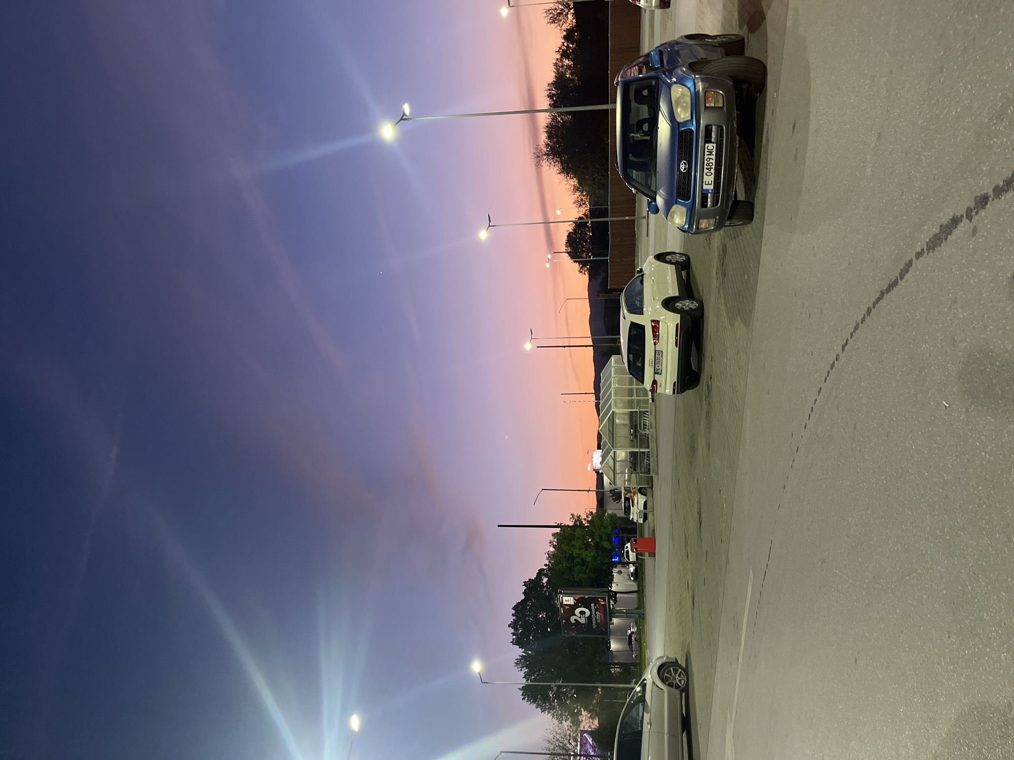 The image captures an outdoor parking lot at dusk in Blagoevgrad, Bulgaria. The original image is rotated 90 degrees counter-clockwise, showing a striking twilight sky that transitions from deep purple-blue at the top to warm orange and pink shades along the horizon.
Several vehicles are parked in the lot, illuminated by numerous bright streetlights that create noticeable lens flares. In the foreground, a blue Toyota RAV4 with the Bulgarian license plate "E 0489 MC" is clearly visible. Behind it, a white Audi displaying the license plate "CK 4699 AB" is partially seen. The ground is asphalt, showing some tire marks.
In the background, trees are silhouetted against the colorful sky. Commercial structures, including shopping cart corrals, suggest the area is associated with a supermarket or retail center. A large vertical sign, partially obscured by trees, features the number "20". While no people are visible, the parked cars and commercial setting imply ongoing shopping or recreational activity during the early evening hours. The scene conveys a calm atmosphere under a beautifully colored sky.