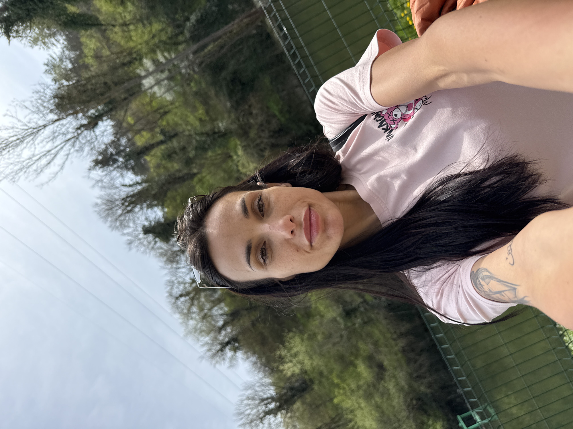 A woman is taking a selfie outdoors. She has long dark hair and is wearing a light pink t-shirt with a cartoon graphic. She is smiling. The background includes green trees and a cloudy sky. There is a wire fence behind her. No specific location in Pleven, Bulgaria can be determined from the image.