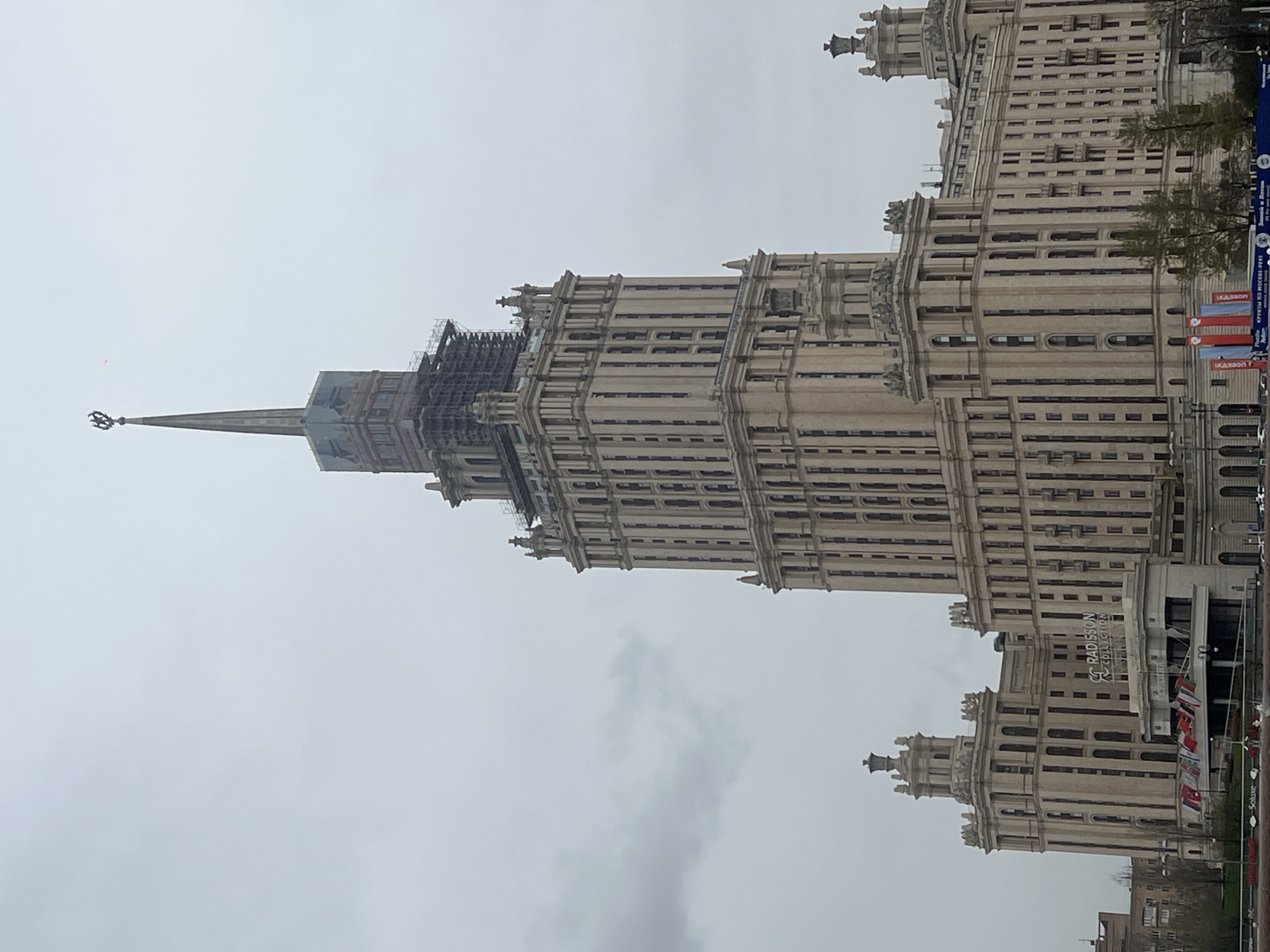 The image shows one of the Seven Sisters skyscrapers in Moscow, Russia, namely the Hotel Ukraina (Radisson Collection Hotel). The building is tall and light-colored, constructed in Stalinist architectural style. The sky is overcast. Part of the building appears to be under renovation, as evidenced by the presence of scaffolding. The Radisson Collection sign is visible, as are several flags.
