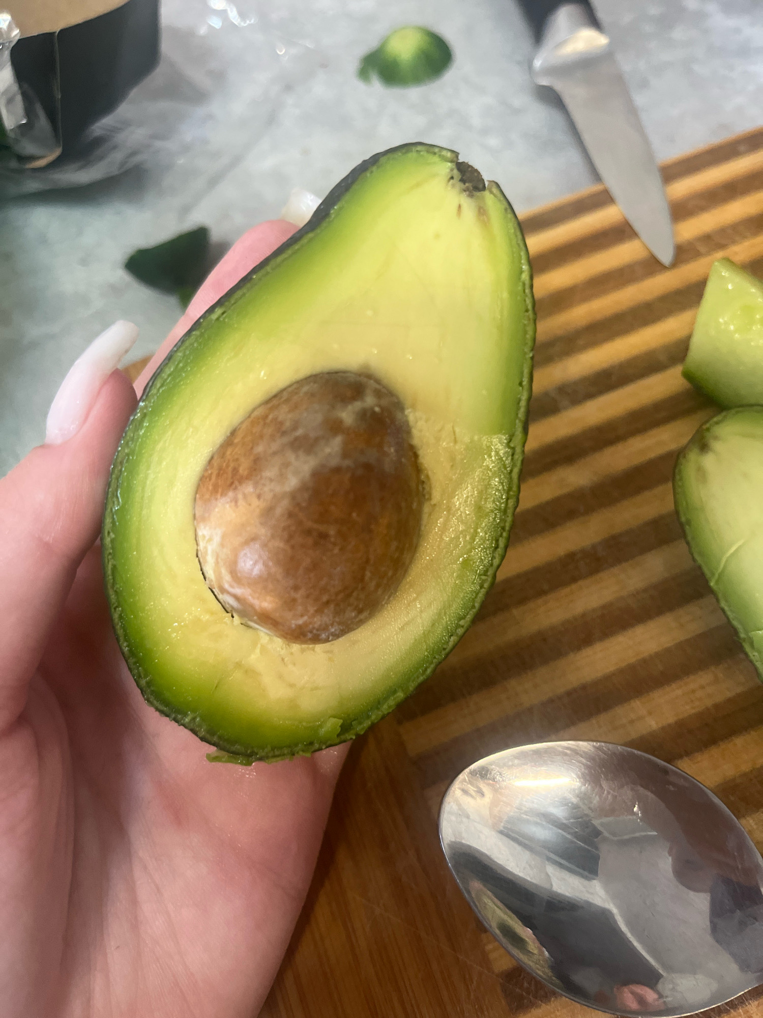 The image depicts a close-up view of a person holding a halved avocado. The avocado is ripe, with a large brown pit. The skin is a vibrant green, and the flesh is creamy yellow. In the background, a wooden cutting board is visible, along with a knife and some chopped pieces of avocado. A silver spoon lies on the cutting board, reflecting the light. The overall scene suggests food preparation. The location is not discernible from the image.