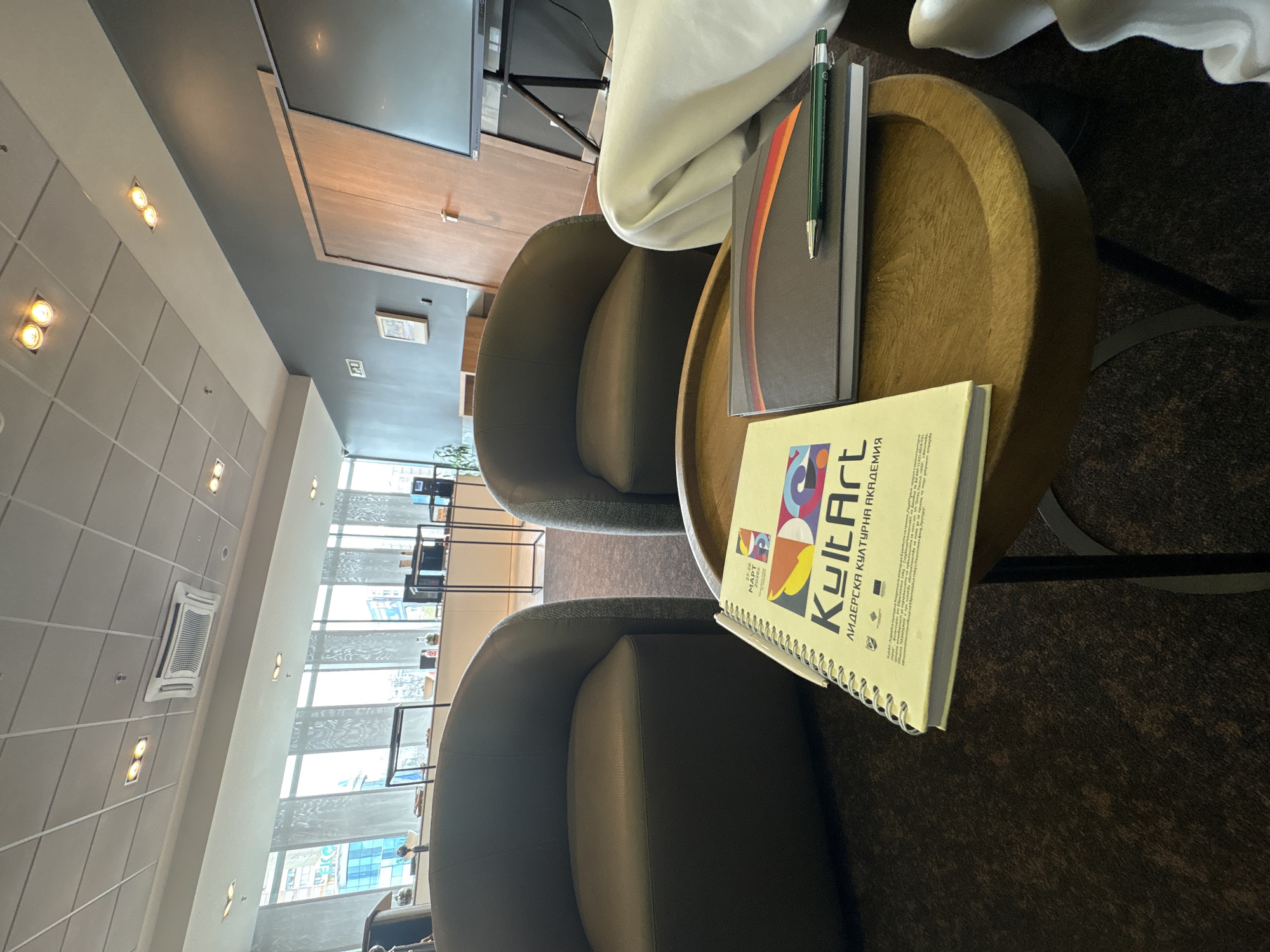 The scene is indoors, likely in a public lounge area or office space. Two armchairs are visible, one on the left side of the frame and one in the foreground, both upholstered in a dark grey or brown fabric. A round wooden table sits between the armchairs. The table holds a notepad with the word "KultArt" and Bulgarian text on its cover, another document with an orange stripe on its edge, and a green pen.
In the background, large windows offer a view of the exterior environment, revealing city buildings, likely in Sofia, Bulgaria. The interior has tiled ceilings with recessed lighting. There is a flat-screen television mounted in the background, and a small picture hanging on the wall.
