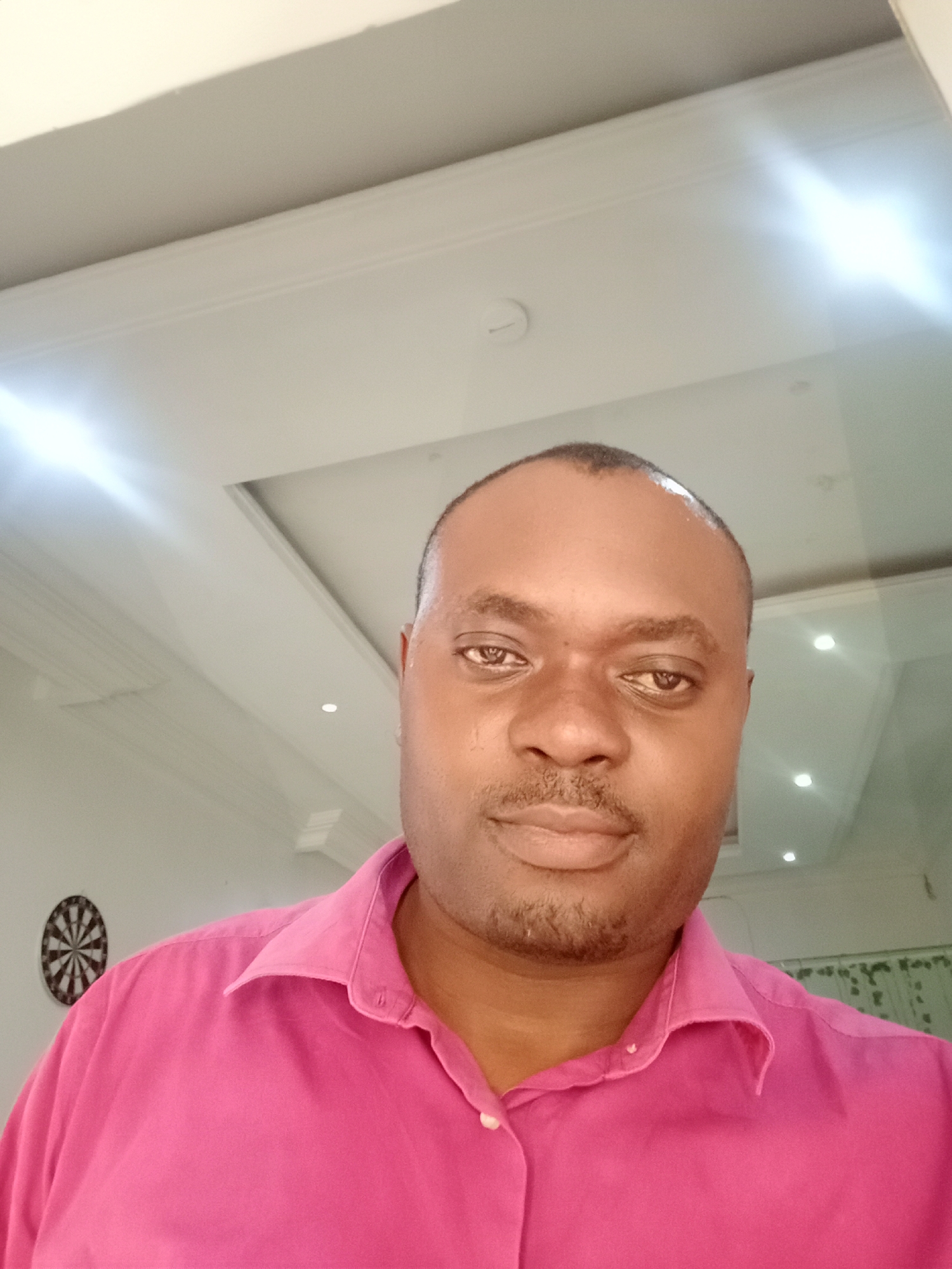 A medium indoor shot of a man in a pink dress shirt. He has dark skin and short dark hair. The background shows a white ceiling with recessed lighting. A dartboard is visible on the left wall. The location is Gwarinpa, Nigeria.