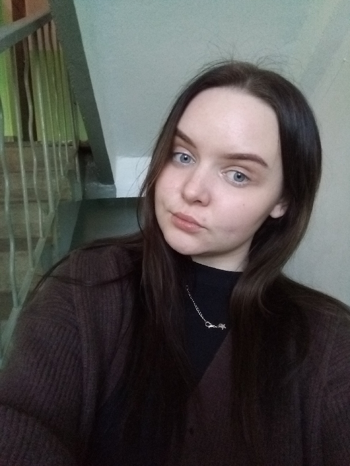 A close-up portrait of a young female subject is presented, set indoors, likely within a residential or public building in the city of Chaikovsky, Russia. The individual has fair skin, blue eyes, long dark brown hair parted down the middle, and light brown eyebrows. She is depicted wearing a black high-necked garment under a dark brown, possibly ribbed, cardigan. A silver-colored chain necklace with a small star-shaped pendant and a visible clasp adorns her neck. Her gaze is directed straight at the camera with a neutral facial expression. The background to the subject's upper left reveals a segment of a stairwell featuring grey metal railings and a light green wall. The remainder of the visible background consists of a plain, light-colored wall.