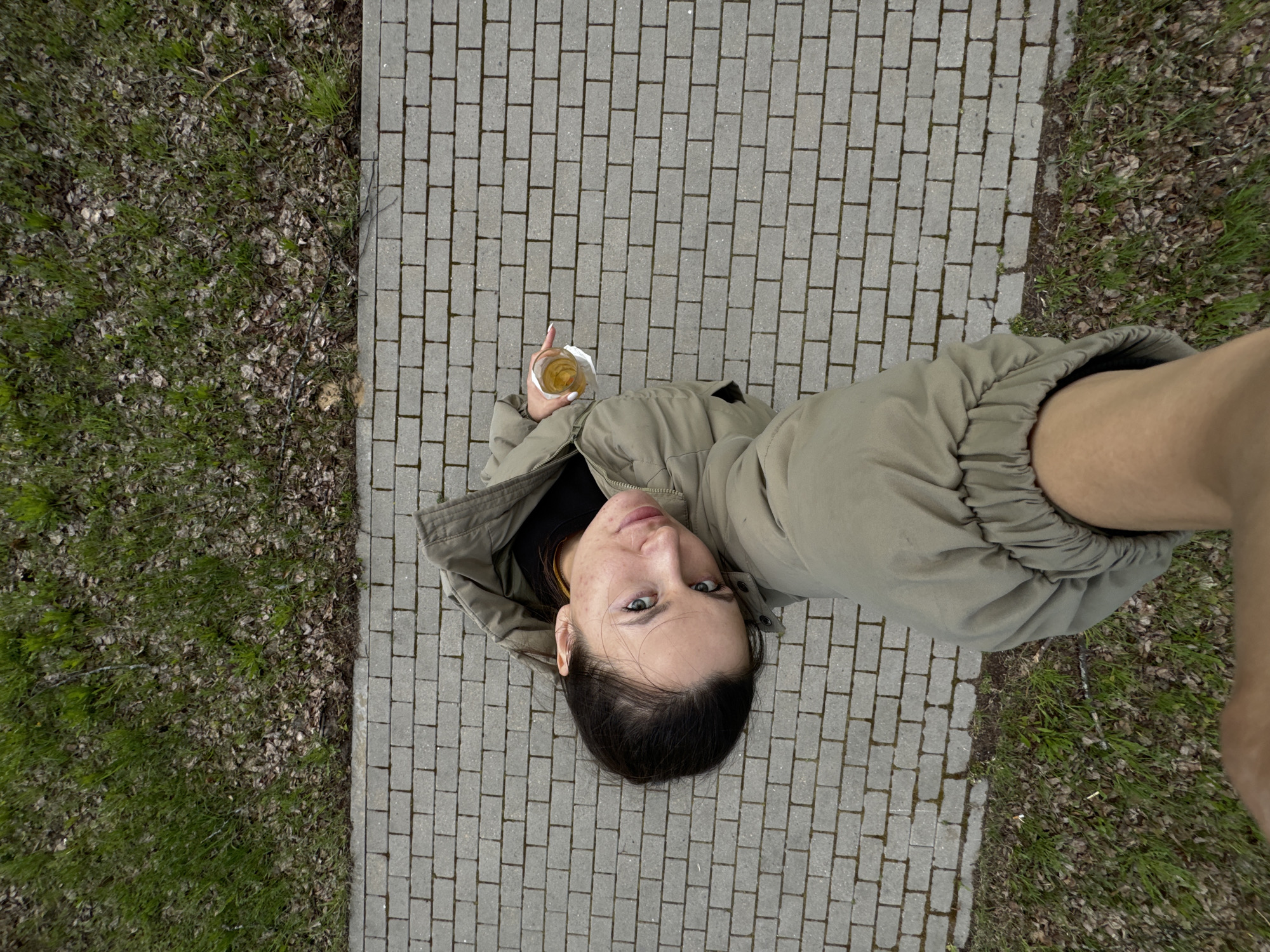 A woman in a light green hooded jacket stands on a gray brick walkway in Красный Маяк, Russia, next to a patch of grass and leaves. She holds a drink in her left hand. The photograph is taken from a high angle, looking down. The woman is looking up at the camera.