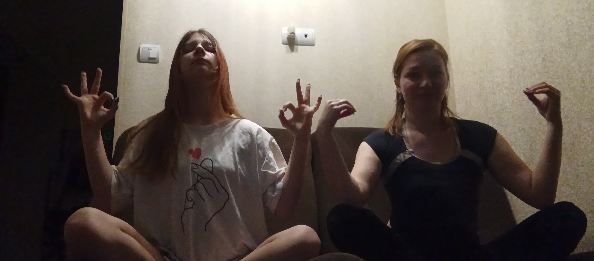 Two women are sitting cross-legged on a brown couch indoors. The woman on the left is fair-skinned with long red-brown hair and wearing a white t-shirt with a design of a hand making a heart. Her left hand is raised in a hand gesture. The woman on the right is fair-skinned with long red-brown hair pulled back and wearing a black t-shirt. Her left hand is raised in a hand gesture. Behind them is a light-colored wall with an electrical outlet and another fixture. The location is Unknown, Unknown.