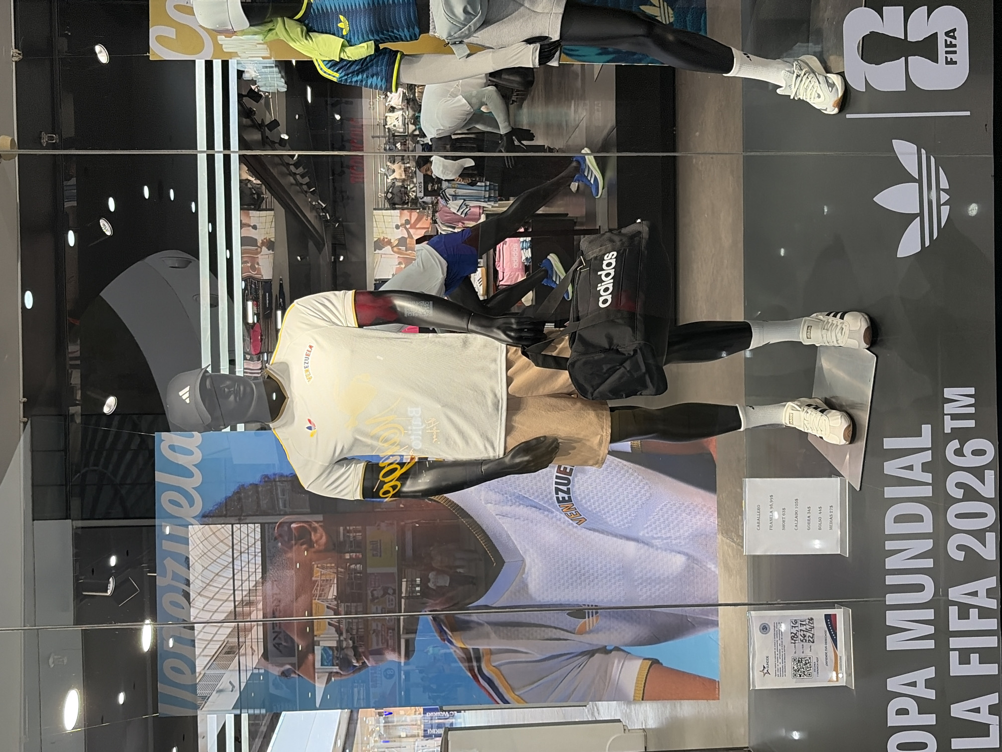 The image shows a storefront display for Adidas athletic wear in Ciudad Guayana, Venezuela. The display features two mannequins dressed in Adidas gear. The main mannequin wears a white and gold Venezuelan national team jersey with a black and gold armband, khaki shorts, and white Adidas sneakers. A black Adidas shoulder bag is slung across its torso. Behind the mannequin is a poster with a picture of a Venezuelan soccer player wearing the same jersey. The poster includes the word "Venezuela." To the right of the mannequin is a sign listing the prices of the clothing in US dollars. The second mannequin in the background wears a blue and yellow Adidas athletic outfit. The text "COPA MUNDIAL LA FIFA 2026 TM" and Adidas logo are displayed on the storefront. The image is taken through glass, reflecting the interior of the store and some of the surrounding area.