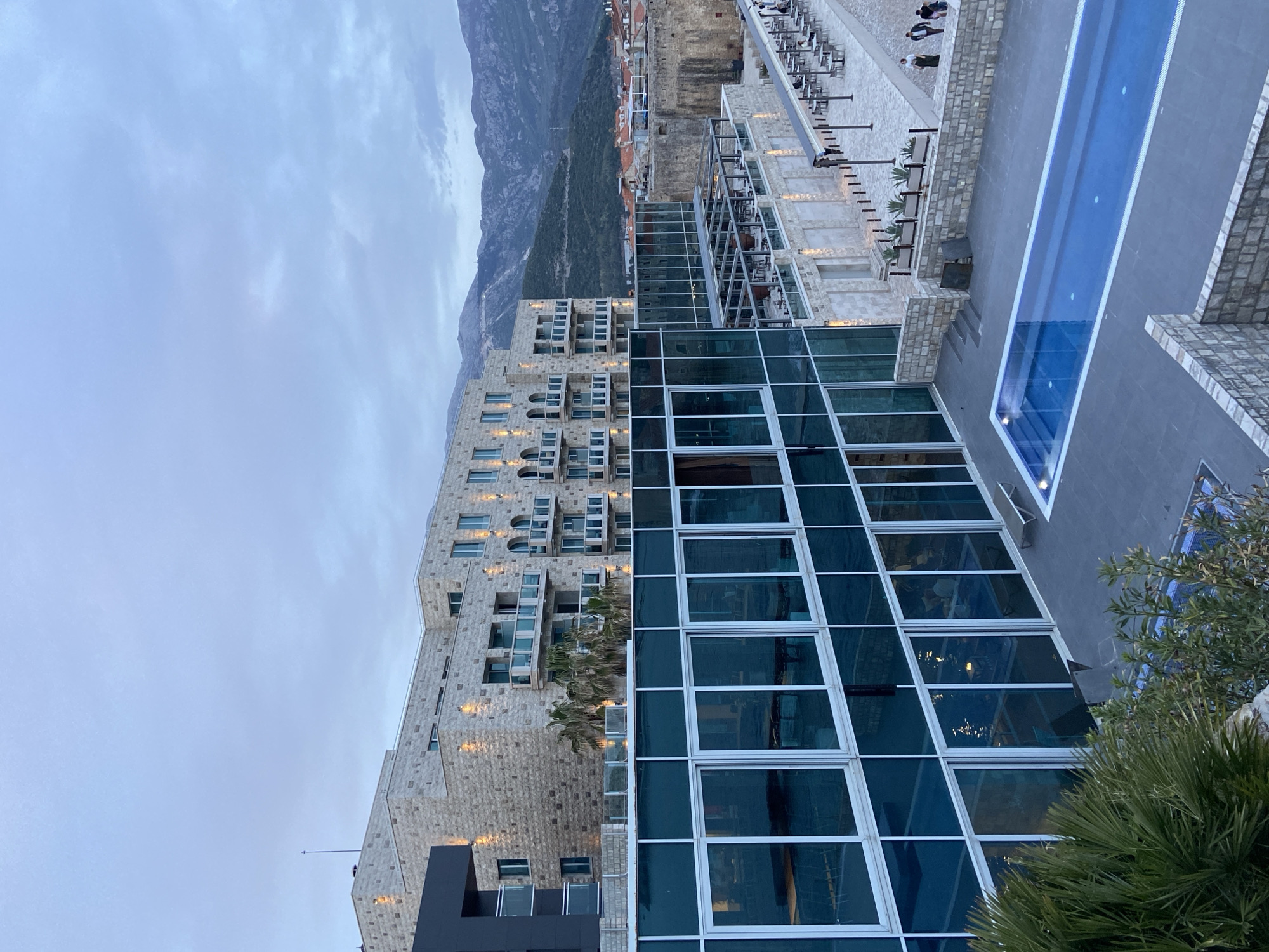 The image shows an elevated view of a modern building complex in Budva, Montenegro, at dusk. The building on the left is a multi-story structure with stone cladding and a facade with numerous windows and balconies. Attached to the building is a large glass-walled structure with a grid-like pattern. Further to the right is a swimming pool. Beyond the buildings is a landscape with mountains in the background. Above, the sky is overcast.