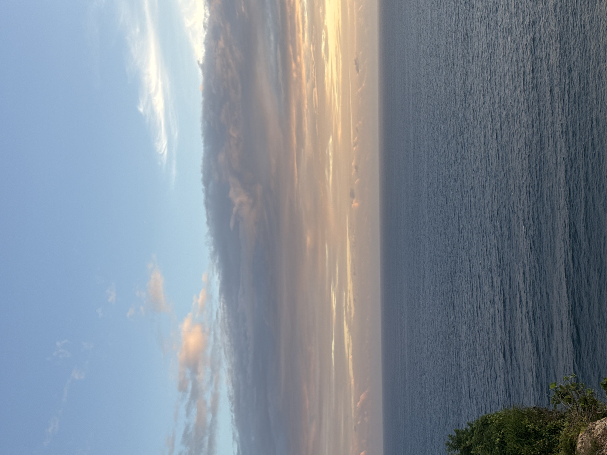 The image shows a view of the ocean meeting the sky, likely taken from an elevated position along the coastline. The sky is a mix of light blue and striated clouds tinged with gold from the sun, suggesting either a sunrise or sunset. The ocean is a dark blue with subtle wave patterns. In the lower right corner, there's a hint of vegetation on a cliff edge. The image is rotated 90 degrees clockwise. The location is Unknown, Unknown.