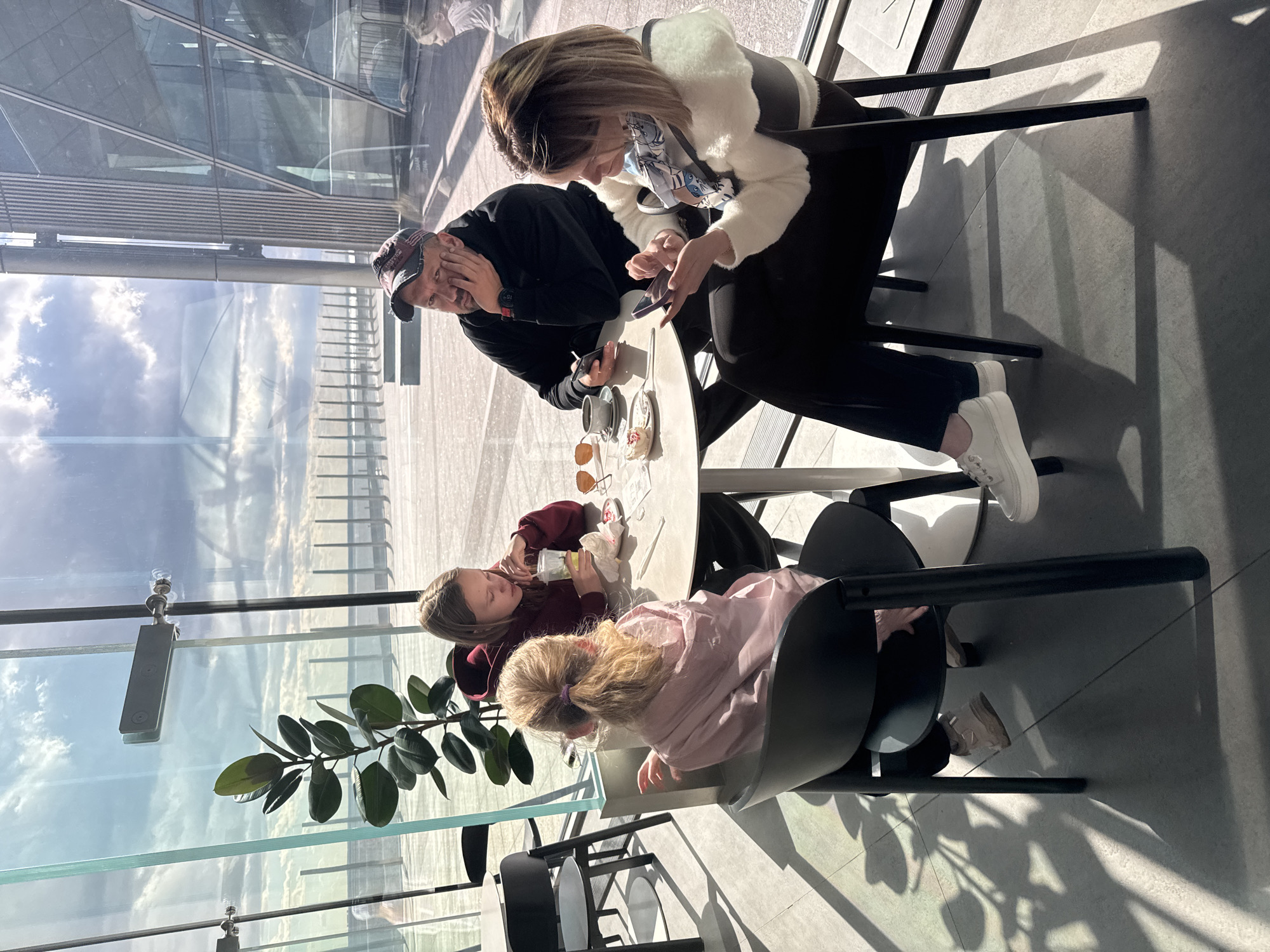 Here is a description of the image:
A family of four is seated at a circular table near a large glass window. The setting appears to be a high-rise building, possibly a restaurant or observation deck, given the expansive view visible through the window. The view includes reflective glass structures and cloudy skies.
The family consists of a man, a woman, and two young girls. The man is wearing a dark jacket and a baseball cap, and he has his hand to his face. The woman is wearing a white jacket with black pants and white sneakers. She is looking at a phone in her hands. The two girls are seated on the opposite side of the table. One wears a burgundy or red top and appears to be drinking from a plastic cup or bottle. The younger girl wears a pink top.
On the table, there are coffee cups, a dessert, sunglasses, and what appears to be a receipt.
The location is likely Saint Petersburg, Russia based on visible reflections of the Lakhta Center (tallest building in Europe) in the windows.