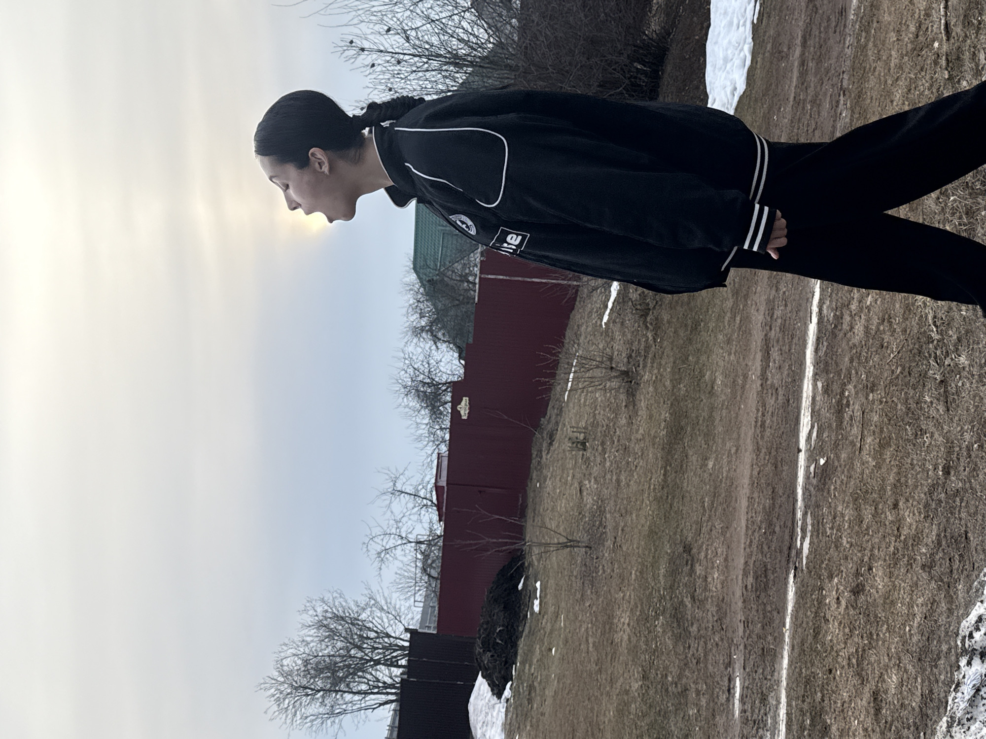 The image shows a woman in a black jacket and black pants standing in an outdoor location in Votkinsk, Russia. The woman is looking towards the sky and has a ponytail. The background includes patches of snow on a grassy area with a red fence and some trees without leaves. The sky is overcast with a hint of sunlight breaking through.