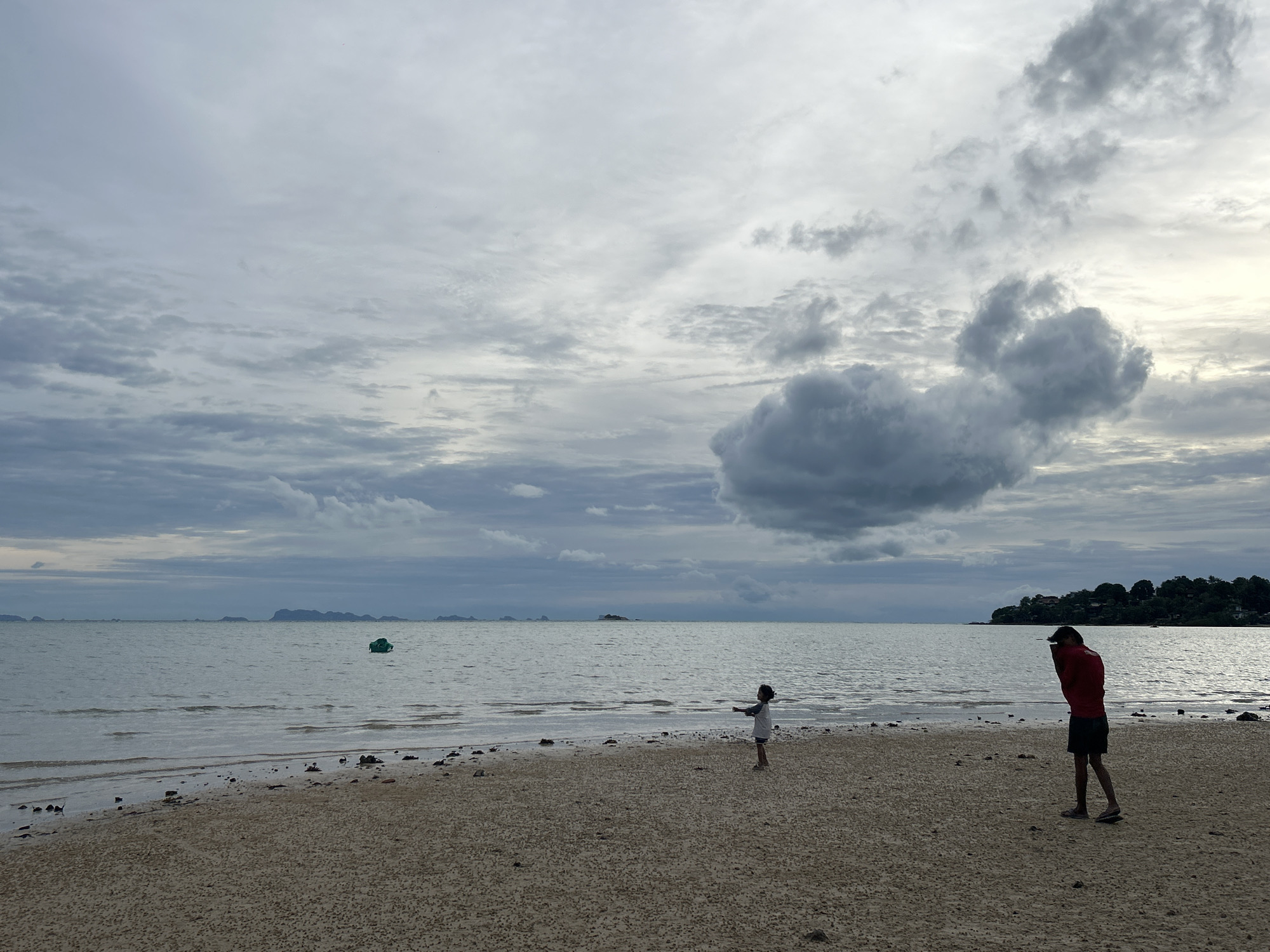 An overcast day at a beach in Ko Pha-ngan District, Thailand. The sand is a light brown with scattered small rocks and debris. The sea is calm with gentle waves. In the distance, several small islands and a green buoy are visible. Two people are on the beach: a child in a white shirt and dark shorts stands facing the sea with arms outstretched, and a person in a red shirt and dark shorts is standing a short distance away, looking down. Trees and some buildings are visible on a distant coastline. The sky is filled with dramatic gray clouds.