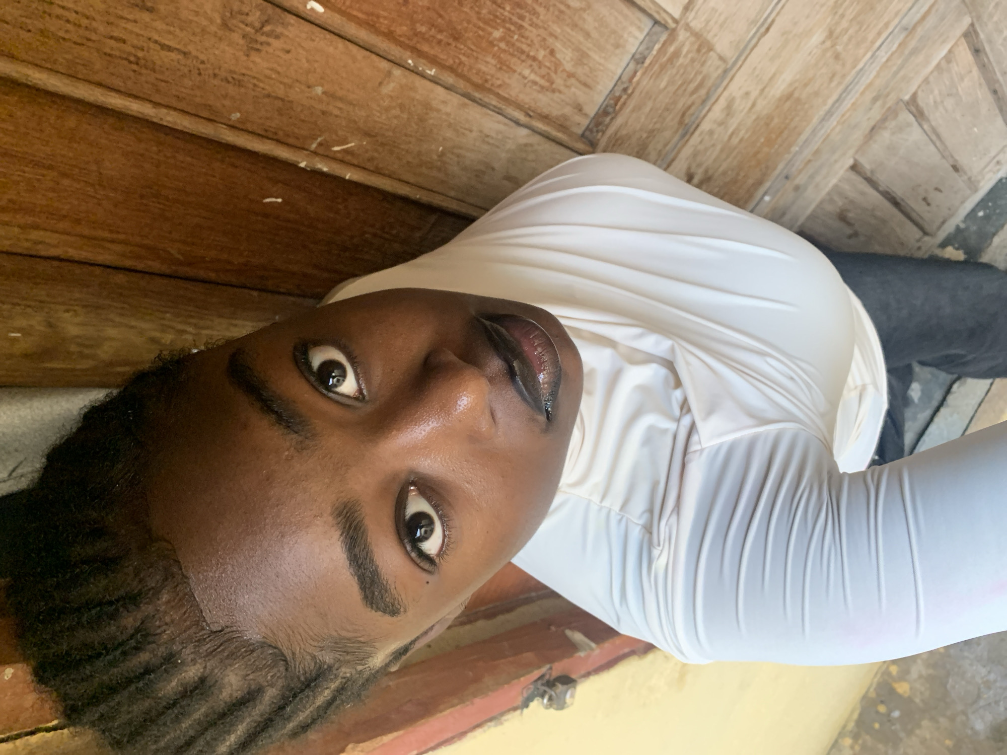A close-up, high-angle shot shows a Black person from the chest up, looking directly at the camera. Their hair is styled in thin braids that are pulled back from their forehead. They are wearing a white, long-sleeved garment with a high neckline, and dark pants. The background consists of a wooden door with vertical planks, and a yellow wall on the left side. The lighting suggests an outdoor or well-lit indoor environment. The scene does not provide any specific details that would allow for identification of a location within Rafin Par, Nigeria.
