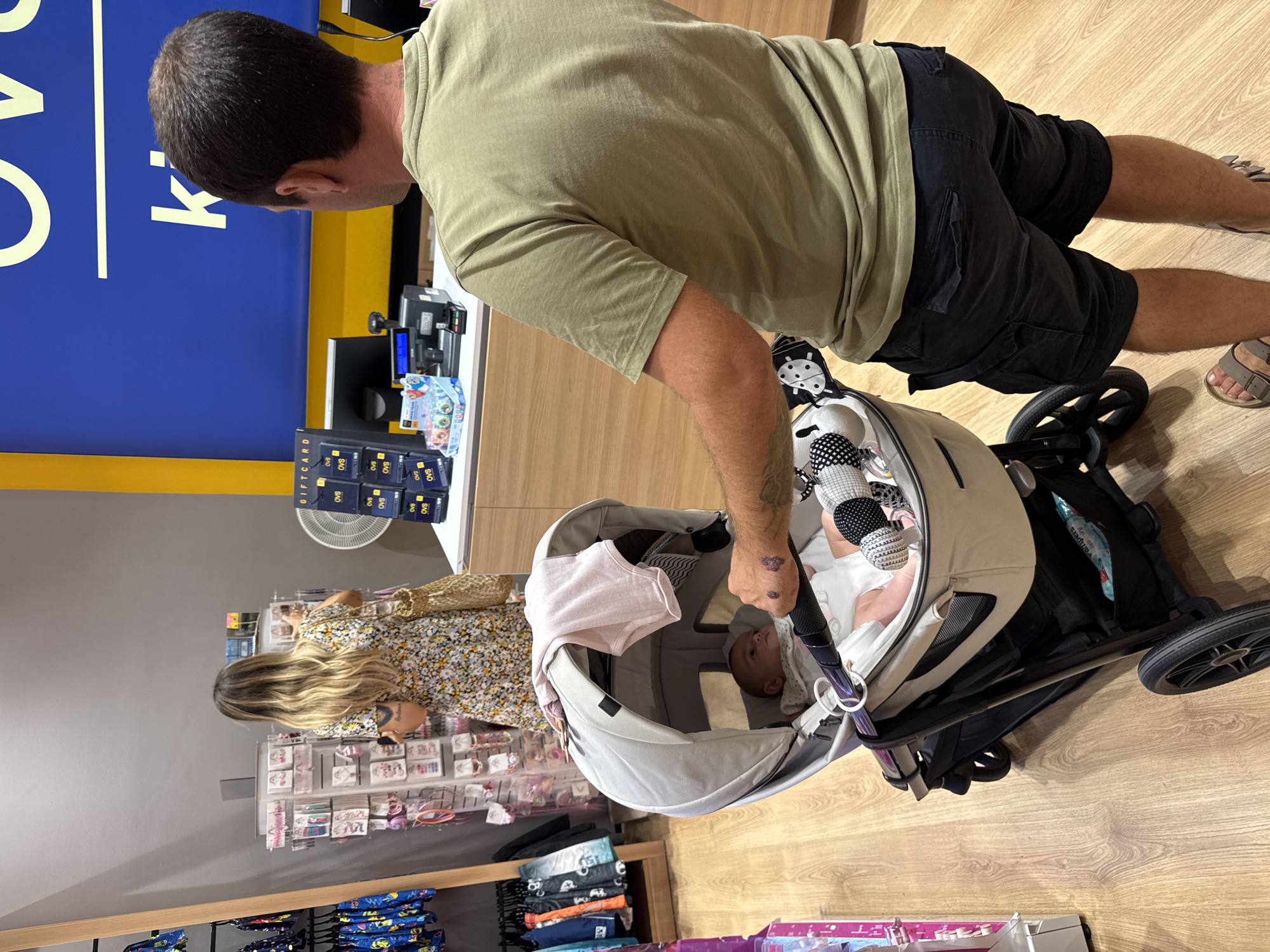 In a retail store in Carini, Italy, a man from the rear is pushing a stroller with a baby inside. He is wearing a green t-shirt and black cargo shorts. A woman with blonde hair is visible in the background, facing away from the camera and browsing a display of merchandise. The store appears to be a clothing or children's goods store, with racks of clothing visible on the left side of the frame. A counter with a cash register is visible in the background. The floor is made of light-colored wood.